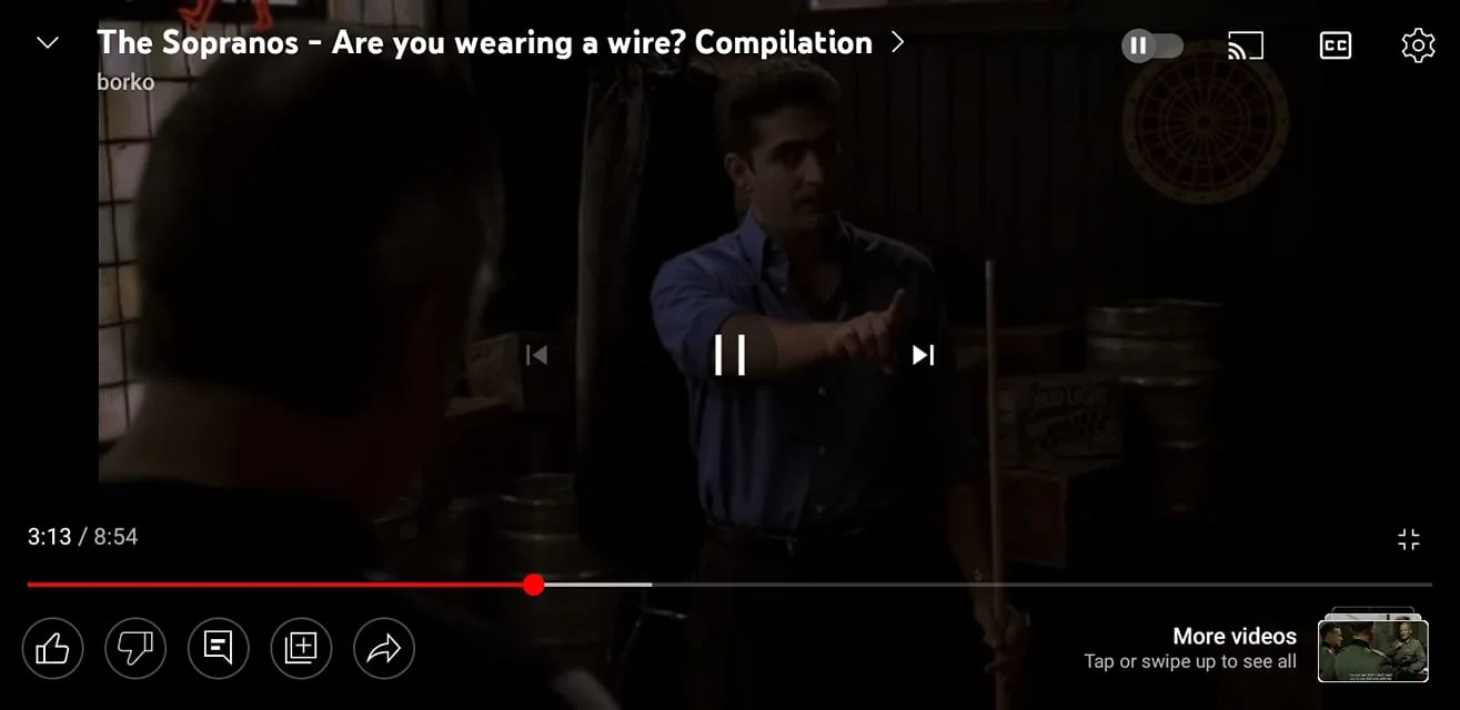 1) Rich description: The image is a still from a YouTube video titled "The Sopranos – Are you wearing a wire? Compilation," uploaded by the channel "borko." The scene is set indoors with dim lighting, suggesting an intimate or secretive conversation typical of "The Sopranos" aesthetic. The focus is on two characters; one is partially visible from the back, while the other stands in a blue shirt, gesturing with a pool cue. The background features a dartboard and stacked barrels, contributing to a casual, possibly bar-like environment. The atmosphere is tense, likely due to the serious nature of the dialogue implied by the title. The man facing forward has a firm expression, suggesting a moment of confrontation or accusation. The video timeline indicates this scene occurs at 3:13 in the compilation, which is 8 minutes and 54 seconds long. The presence of typical YouTube controls like play, pause, and captions suggests an online video viewing interface.
2) On-screen text block:
- Title: "The Sopranos – Are you wearing a wire? Compilation"
- Channel / profile: "borko"
- Site / app: "YouTube"