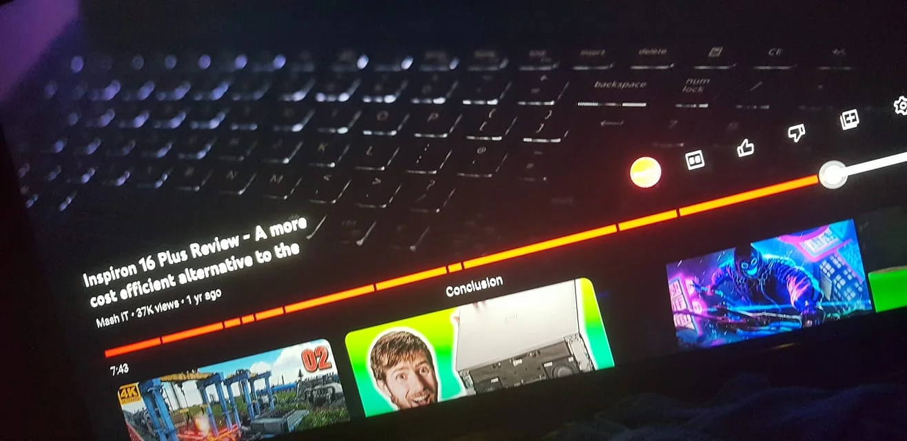 The image is a screenshot from a YouTube video. It features a review titled "Inspiron 16 Plus Review - A more cost efficient alternative to the," presented by the channel "Mash IT." The video has accumulated 37K views and was posted a year ago. The progress bar indicates a position at 7:48, with thumbnails from different parts of the video visible at the bottom. The background shows a blurred image of a laptop keyboard, suggesting the tech-focused theme of the content. The interface elements include icons for like, dislike, and share, and the overall setting appears to be a dimly lit indoor environment, likely to emphasize the screen. This setup creates a focused atmosphere typical of tech review content.