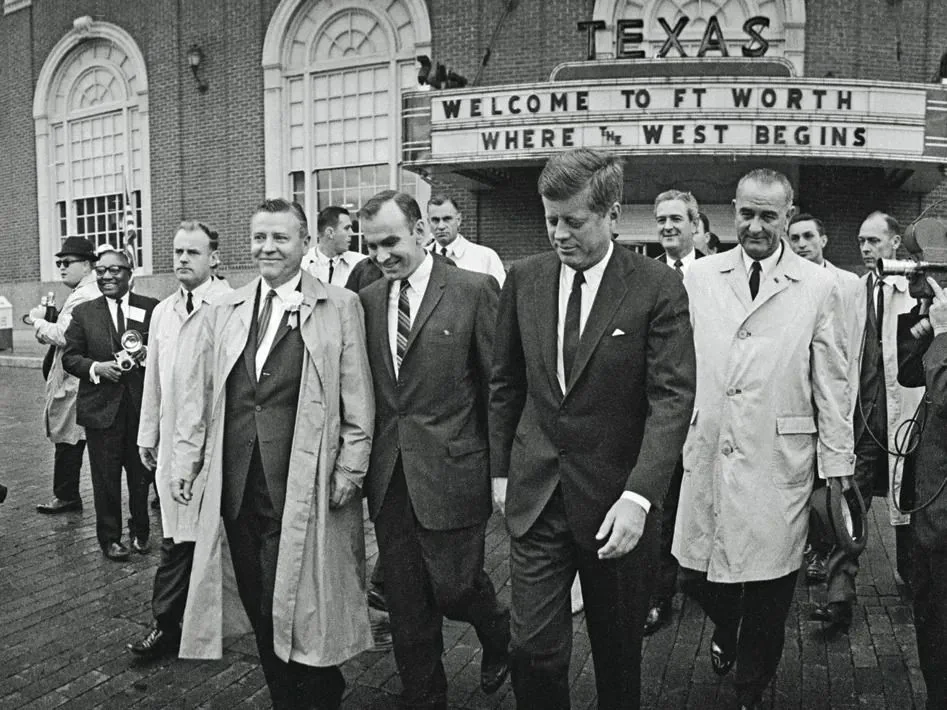 In this historical black-and-white photograph, a group of men is seen walking together outside with a building marquee in the background reading "WELCOME TO FT WORTH WHERE THE WEST BEGINS." Prominently, JFK and LBJ are among them, based on the hint. JFK is wearing a dark suit and tie, exuding a formal and composed demeanor. Beside him, a man identified from the hint as LBJ is dressed in a lighter trench coat, walking alongside others who appear to be officials or aides.
The men are gathered on a wet cobblestone street, suggesting rainy conditions. Several other individuals, some holding cameras, follow closely, indicating a media presence capturing the moment. The setting appears to be a welcoming event, likely during a political visit or campaign stop in Fort Worth, Texas. The mood is professional and cordial, with the men engaged in conversation or focused on their surroundings. The architecture of the building, with its arched windows, adds a classic backdrop to the scene.
