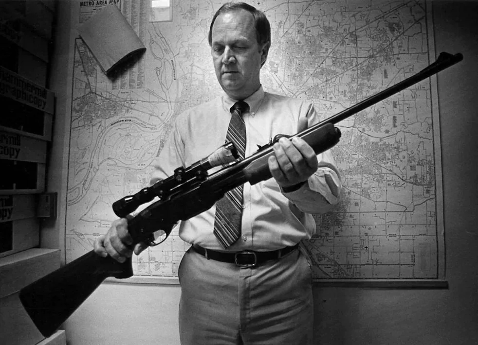 The black and white image depicts a man in a business office environment, examining a rifle with a scope. He is dressed in a light-colored shirt and patterned tie, and his demeanor is serious and focused as he holds the firearm. The background features a large map labeled "Metro Area Map," suggesting an urban setting, possibly within a police or investigative department. The room appears organized, with shelves containing boxes and documents. The lighting is soft, with a professional atmosphere typical of an office. The man's posture is attentive, indicating he is either inspecting or demonstrating the weapon. The photograph captures a moment that likely pertains to law enforcement activities, with the man possibly being an official or expert in firearms. The lack of color emphasizes the seriousness and formality of the scene.