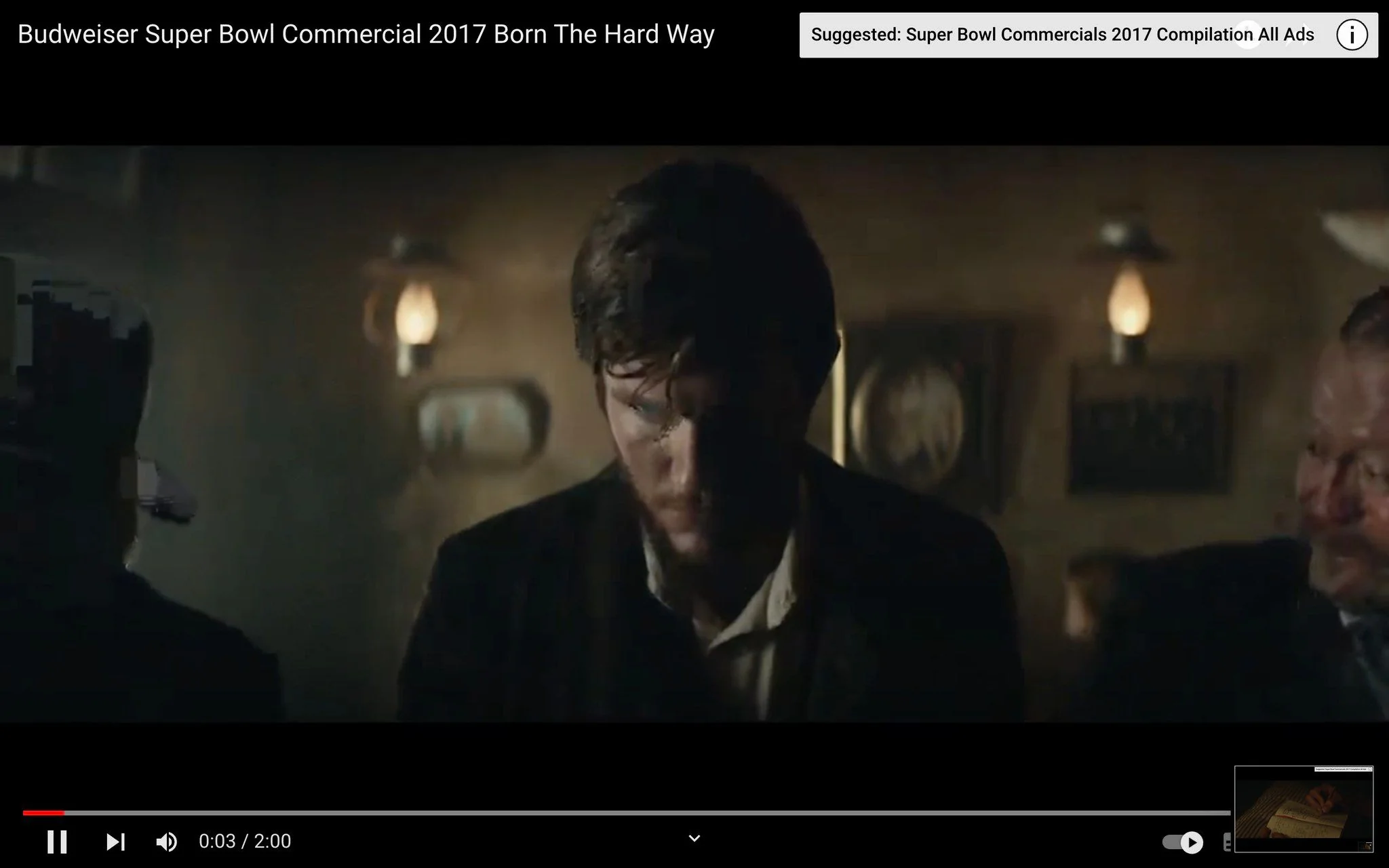 The video frame is from the Budweiser Super Bowl Commercial 2017 titled "Born The Hard Way." It depicts a dimly lit, historical setting with a dramatic atmosphere. The scene captures a man in the center, who appears to be in a contemplative or somber mood. His attire suggests a period piece, possibly from the late 19th or early 20th century, featuring a dark, rugged suit and a white shirt. The lighting is soft and subdued, casting shadows and enhancing the nostalgic ambiance. The background shows a rustic interior with wall-mounted lamps and blurred decorative elements, further enhancing the vintage feel. The overall mood of the frame is one of introspection and resilience, aligning with the commercial's theme of overcoming adversity. This frame is part of a narrative that explores themes of ambition and determination, often associated with the brand's storied history.
- Title: "Budweiser Super Bowl Commercial 2017 Born The Hard Way"
- Channel / profile: "Suggested: Super Bowl Commercials 2017 Compilation All Ads"
- Site / app: "YouTube"