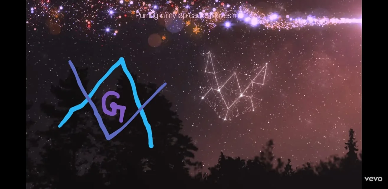 The image features a night sky filled with stars and constellations set against a silhouette of trees in the foreground. The sky is beautifully illustrated with a starry band across the top, resembling the Milky Way, with a mix of purples, blues, and whites. Among the stars, there is an artistic representation of a constellation drawn with lines connecting various stars, forming geometric shapes. On the left side, there is a stylized drawing with the letter "G" in purple, surrounded by intersecting blue lines forming a diamond shape. The words "Purring in my lap cause it loves m" are visible at the top, suggesting a lyrical or poetic context. The logo for "vevo" appears in the bottom right, indicating the platform where this image might be featured or referenced. The overall mood is serene and mystical, emphasizing the beauty of the cosmos.