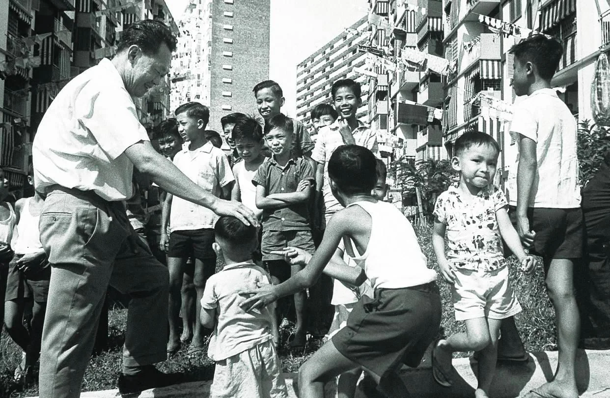 In the black-and-white image, Lee Kuan Yew is depicted engaging with a group of children in an outdoor setting. The scene takes place in what appears to be a residential area, with tall apartment buildings in the background. Lee Kuan Yew is seen bending slightly as he interacts with the children, gently patting one child on the head. The children, dressed in casual summer clothing like shorts and T-shirts, are gathered around him, some smiling and others appearing curious and attentive. The mood is lively and positive, suggesting a community event or visit. The lighting indicates it is daytime, casting shadows on the ground, and the overall atmosphere is friendly and relaxed. Lee Kuan Yew's presence and interaction suggest his role as a leader connecting with the people. The image captures a moment of warmth and connection between him and the local community.