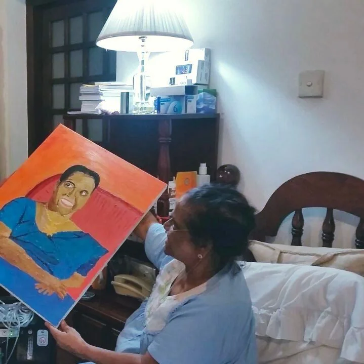 The image depicts Milina Sumathipala in a cozy indoor setting, possibly her home. She is holding a colorful portrait painting, which appears to be quite dear to her, as suggested by the hint. The portrait is vibrant, featuring a person in a blue shirt against a red and orange background. Milina is seated on a bed with white pillows, and the room is softly illuminated by a lamp on a nearby bedside table. The table also holds several items like books and small boxes, adding a lived-in feel to the scene. Her posture is relaxed and she seems to be admiring or contemplating the painting, suggesting a personal connection. The lighting in the room is warm, contributing to the intimate and peaceful atmosphere. The focus on the portrait and Milina’s interaction with it highlights its significance as her most prized possession.