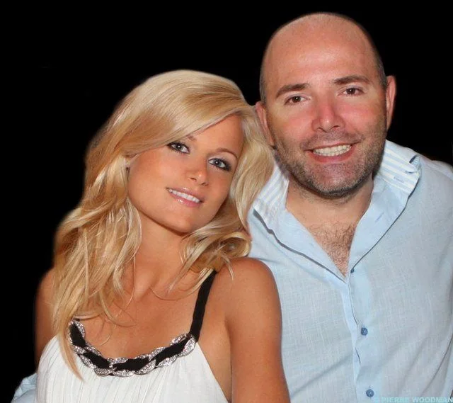 In this image, two people are posing together against a plain dark background. The man, identified as Pierre Woodman based on the hint provided, has a cheerful expression. He is wearing a light blue, button-up shirt with a slight open collar, and his head is shaved. The woman beside him has long, wavy blonde hair and is wearing a white top with black straps and decorative elements along the neckline. She is leaning slightly towards Pierre, creating a sense of closeness. Both individuals appear relaxed and are smiling warmly, suggesting a friendly or familiar relationship. The lighting is soft and even, highlighting their features against the dark backdrop. The atmosphere of the image conveys a candid and informal moment captured between the two.