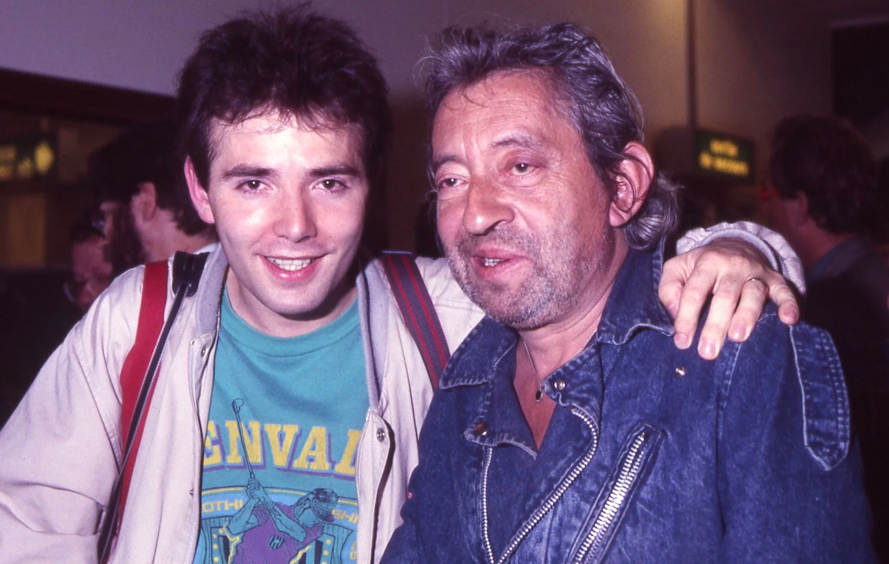In the image, two men are posing together indoors, possibly at an airport, given the background signage. The individual on the left, identified as Pierre Woodman, is wearing a casual outfit consisting of a light jacket over a green t-shirt featuring a graphic design and text that reads "NEVAL." He has a backpack strap over his shoulder, suggesting he might be traveling. His expression is cheerful, and he appears relaxed and friendly. The man on the right, wearing a denim jacket with a zipper and a rugged, casual style, has an arm around Woodman in a gesture of camaraderie. His expression is relaxed and slightly amused. The lighting is bright and typical of an indoor public space, highlighting their faces clearly. The overall mood of the image is informal and friendly, capturing a candid moment of friendship or acquaintance.