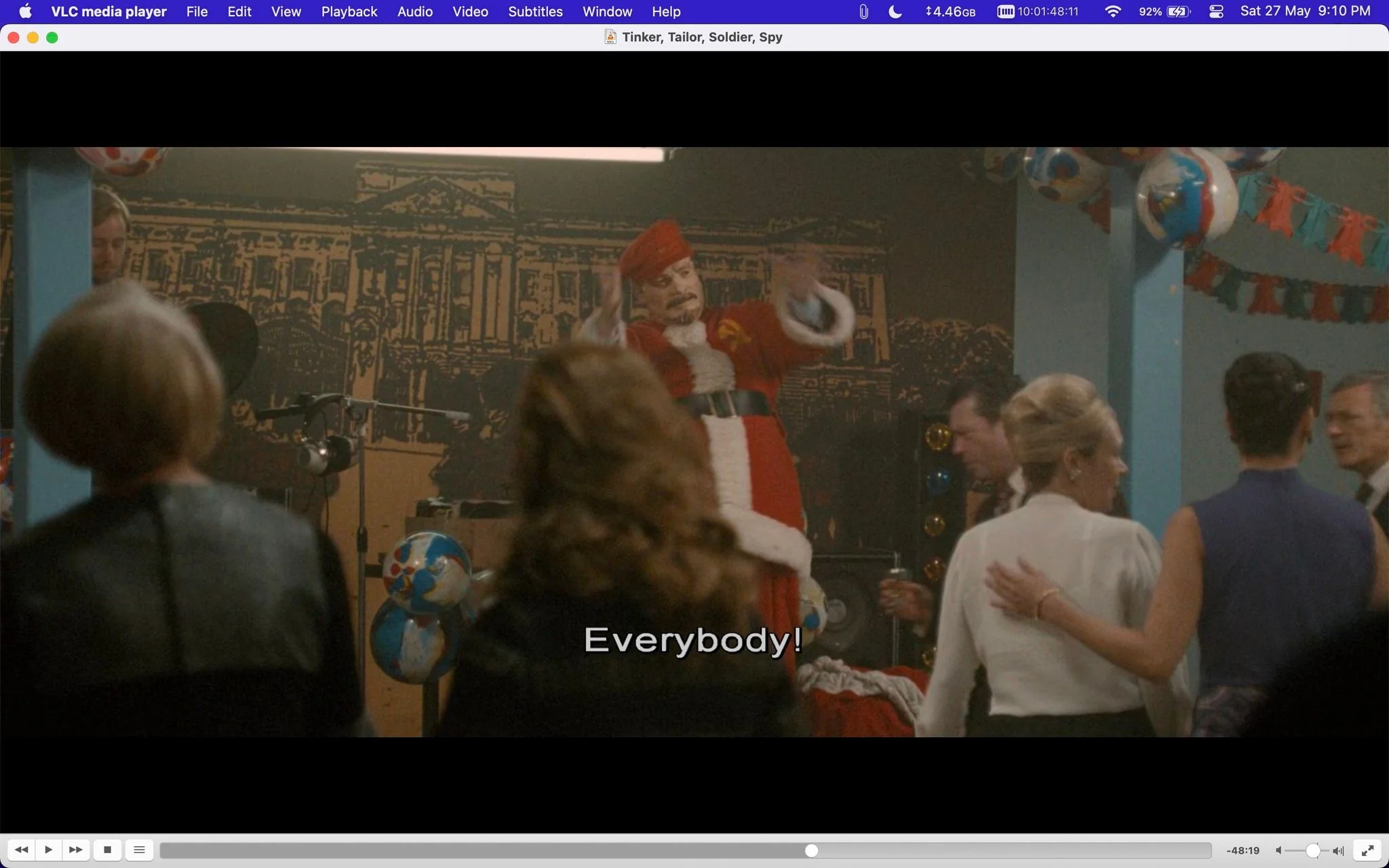 The image is a frame from the film "Tinker, Tailor, Soldier, Spy," displayed in a VLC media player window. The scene takes place indoors, likely at a party or gathering. In the middle of the frame, a man dressed in a red and white Santa-like outfit, complete with a hat and black belt, is animatedly gesturing as if addressing a crowd. The audience is partially visible, with several people standing and facing the stage, indicating they are engaged in the performance. Decorations such as balloons and streamers are visible, suggesting a festive atmosphere. The lighting is warm, and the background features a large mural or painting depicting a classical building. The subtitle at the bottom reads "Everybody!" indicating that the speaker is likely calling for attention or participation from the audience. The scene captures a lively and celebratory moment within the narrative of the film.
- Title: "Tinker, Tailor, Soldier, Spy"
- Site / app: "VLC media player"
- Captions / subtitles: "Everybody!"