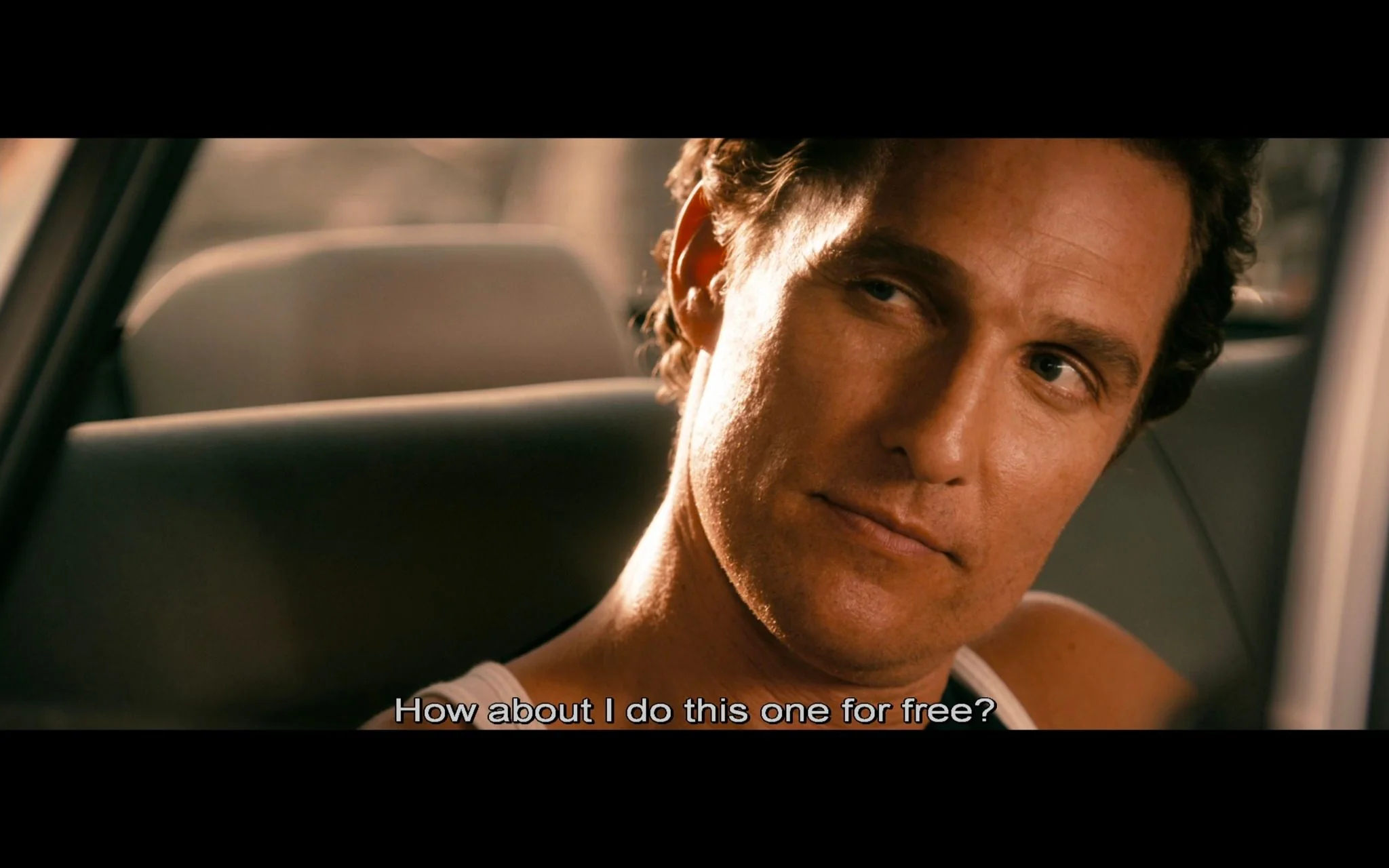 The image is a scene from the movie "The Lincoln Lawyer" featuring a character played by Matthew McConaughey. The setting is inside a car during the daytime, with sunlight casting a warm glow through the window. Matthew's character is seated, wearing a sleeveless shirt, and is looking slightly to the side with a contemplative expression. The focus is on his face, capturing a moment of dialogue, as indicated by the subtitle at the bottom that reads, "How about I do this one for free?" The close-up shot emphasizes his expression and demeanor, suggesting a moment of decision or negotiation. The background is softly blurred, keeping the attention on the character and his expression.