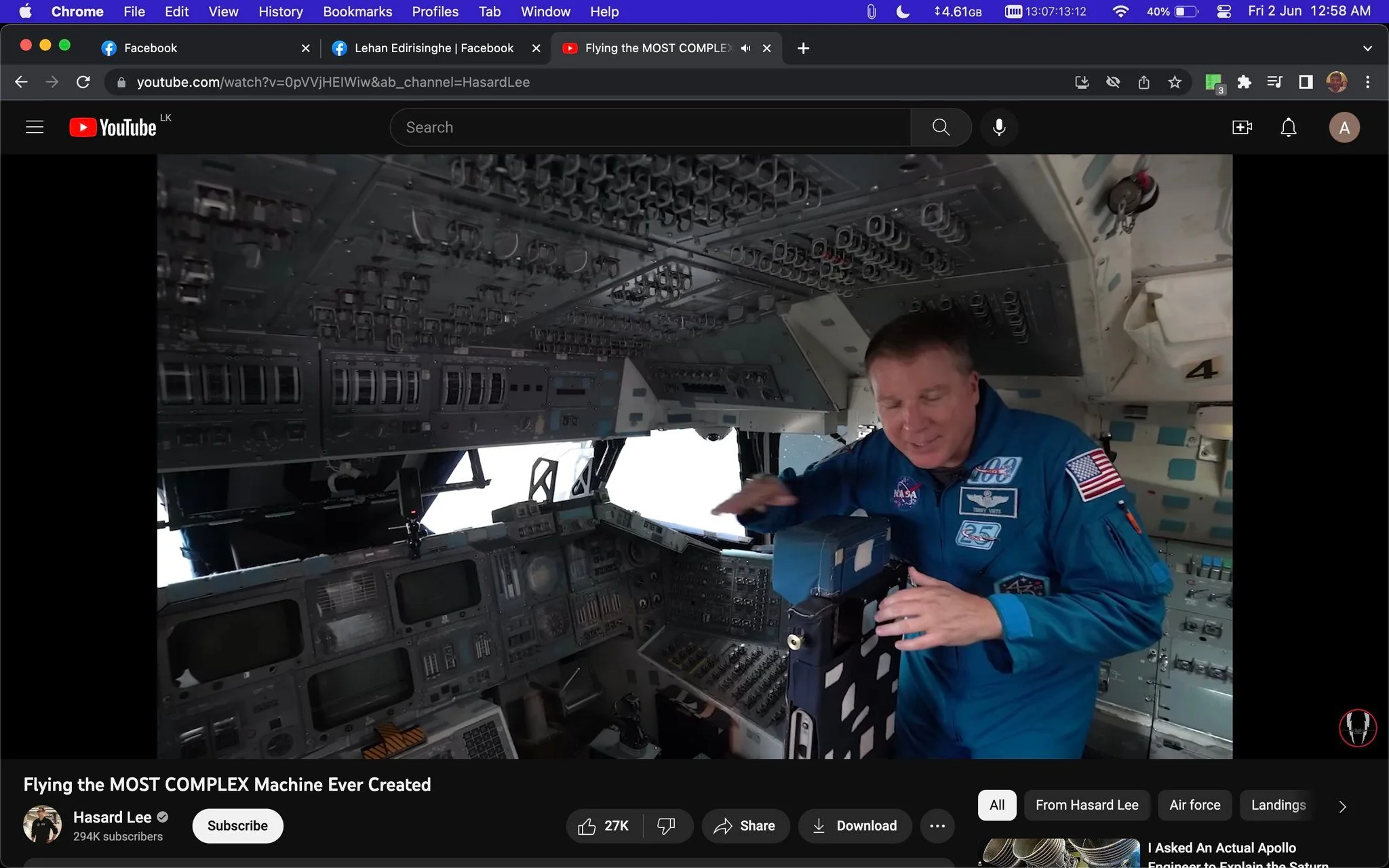 The image is a screenshot from a YouTube video titled "Flying the MOST COMPLEX Machine Ever Created" on the channel "Hasard Lee." The scene is set inside a spacecraft cockpit, filled with numerous dials, switches, and screens, indicative of a highly technical environment. A person wearing a blue astronaut jumpsuit with NASA patches is present, gesturing towards the equipment. The suit features an American flag patch on the shoulder and a patch celebrating 25 years. The lighting is even, highlighting the complexity of the cockpit's interior. The focal point is the interaction between the individual and the sophisticated machinery, emphasizing the intricacy and advanced technology of the spacecraft.