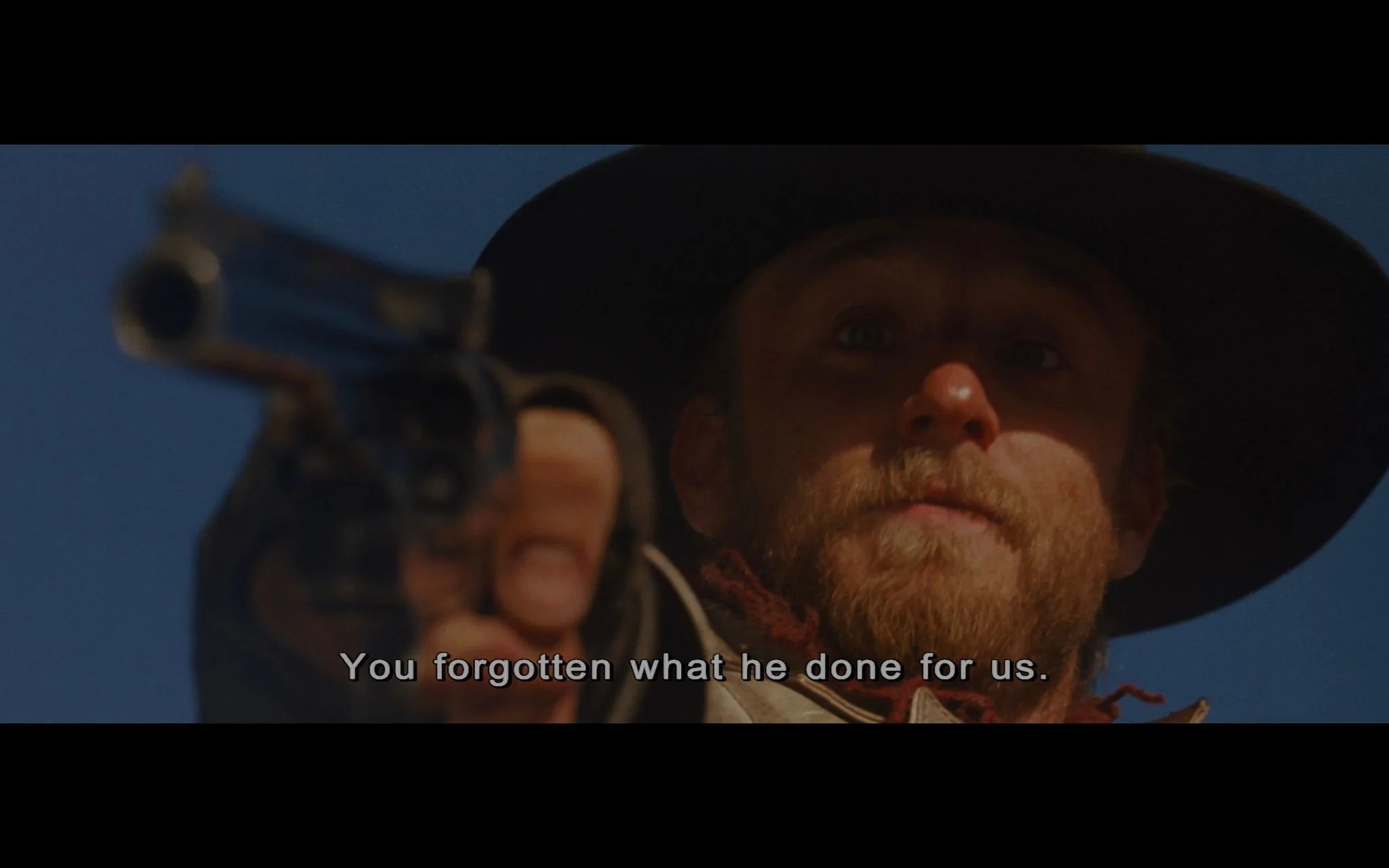 The image is a tense video frame from the film "The Assassination of Jesse James by the Coward Robert Ford." The scene captures a close-up of a man with a beard and a serious expression, wearing a wide-brimmed black hat, pointing a gun directly towards the camera. His eyes are focused, and the lighting casts a shadow over part of his face, enhancing the dramatic intensity. The background is a clear blue sky, suggesting an outdoor setting. The man's rugged attire and the western style hint at the historical context of the movie. The subtitle reads, "You forgotten what he done for us," indicating a moment of confrontation or reflection on past actions. The framing and lighting emphasize the gravity of the situation, typical of a Western drama.