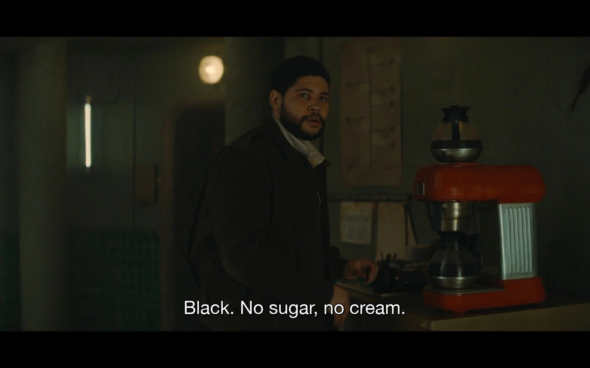The image is a dimly lit interior scene featuring a man standing next to a coffee machine. The setting appears to be a somewhat cozy but dimly lit room, with muted green walls and soft lighting that casts gentle shadows. The man is wearing a dark jacket over a lighter shirt, suggesting a casual or work environment. He is looking towards the camera, possibly engaging in conversation, with a serious yet relaxed expression. The coffee machine in the foreground is red and silver, adding a pop of color to the otherwise subdued palette of the room. It is positioned on a counter, and a coffee pot is visible on top. The atmosphere feels intimate and perhaps slightly tense, suggested by the straightforward nature of the caption: "Black. No sugar, no cream." This indicates a preference or instruction regarding coffee preparation, highlighting a small but telling character detail.