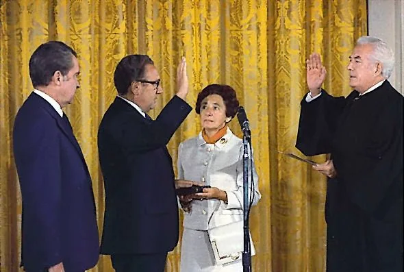 ```
In this image, Henry Kissinger is taking an oath during a formal swearing-in ceremony. The setting appears to be an ornate room, possibly a government building, with a prominent gold curtain in the background. The composition is centered around four individuals: Henry Kissinger, a woman holding a bible, a judge administering the oath, and another man in the foreground.
Henry Kissinger, dressed in a dark suit and glasses, is raising his right hand. The woman beside him is holding a bible close, suggesting her role in the oath-taking process. The judge, also in a dark robe, mirrors Kissinger's gesture by raising his right hand, a traditional aspect of such ceremonies. The mood is solemn and formal, with all individuals appearing serious and focused on the proceedings. The lighting is bright, illuminating the participants and highlighting the rich textures and colors of their attire and the setting. The moment captures a significant event in a political context, emphasizing the importance of the ceremony.