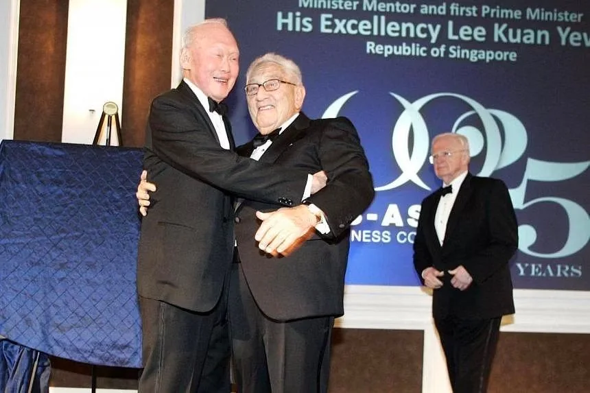 The image captures a formal event featuring Lee Kuan Yew and Henry Kissinger. Both are dressed in elegant black tuxedos, indicative of a high-profile evening occasion. Lee Kuan Yew, with a bright smile, is warmly embracing Henry Kissinger, who also appears cheerful. This moment is set against a backdrop featuring celebratory text that reads "Minister Mentor and first Prime Minister His Excellency Lee Kuan Yew Republic of Singapore," suggesting a tribute or anniversary event.
The mood is warm and cordial, with both men appearing relaxed and content in each other's company. The lighting is bright, accentuating the formality of the attire and the significance of the occasion. A third man in a tuxedo stands in the background, observing the interaction. The venue's decor, with a blue cloth draped over a stand and elegantly designed pillars, enhances the sophistication of the scene.