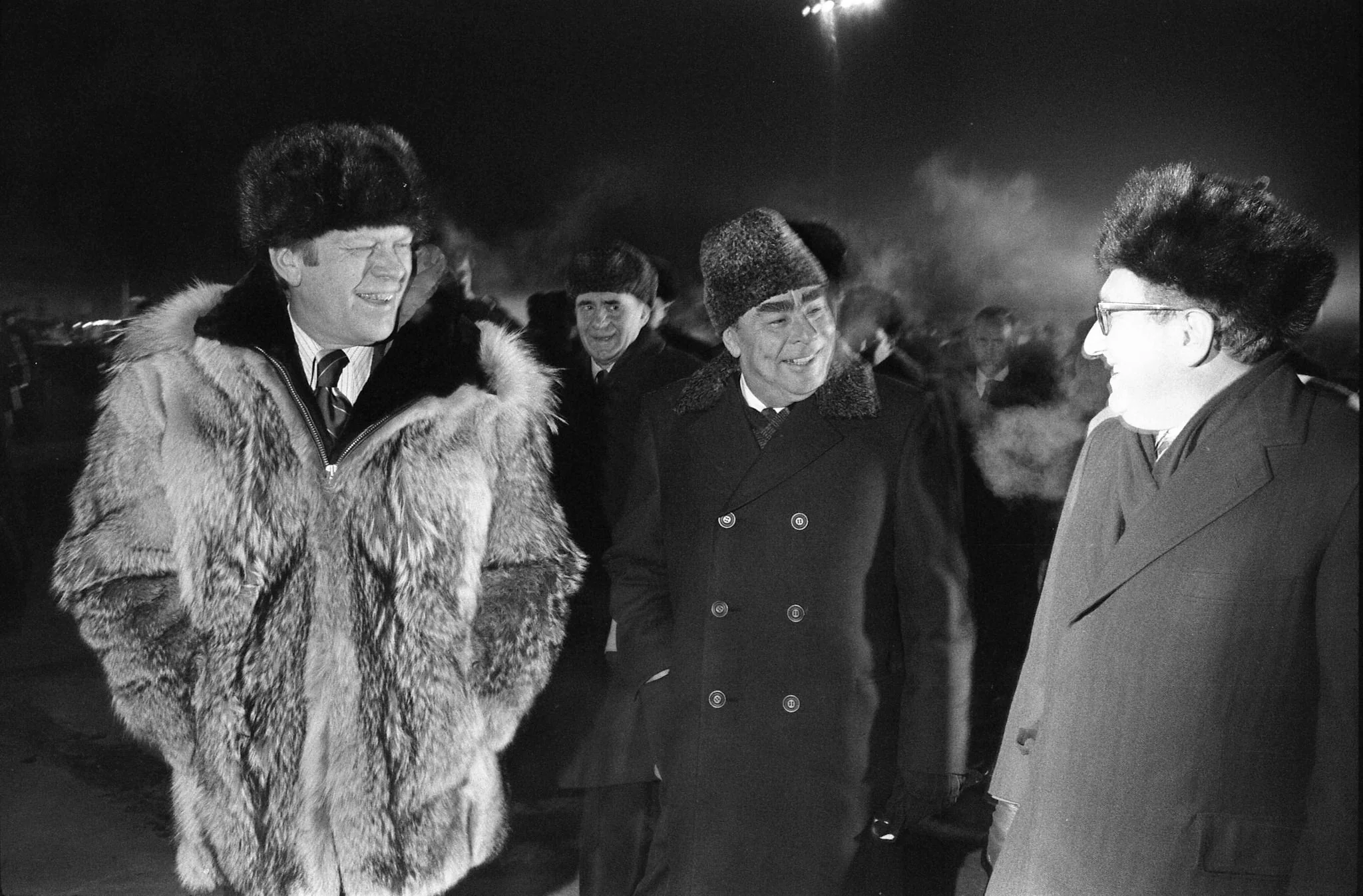 In this black-and-white photograph, the scene takes place outdoors at night in a cold environment, suggested by the visible breath of the individuals and their attire. Three men are walking together, dressed in heavy winter coats and fur hats. The man on the right is identified as Henry Kissinger. He is wearing a dark overcoat and a fur hat, with glasses, and appears to be engaged in conversation with the men beside him. The man in the center is wearing a double-breasted coat and a ushanka-style hat, while the man on the left is dressed in a voluminous fur coat and a similar fur hat. Their expressions are jovial, suggesting a cordial interaction. The background is dimly lit, with additional figures visible behind them, hinting at a formal or diplomatic gathering. The overall mood is one of warmth and camaraderie amidst the cold setting.