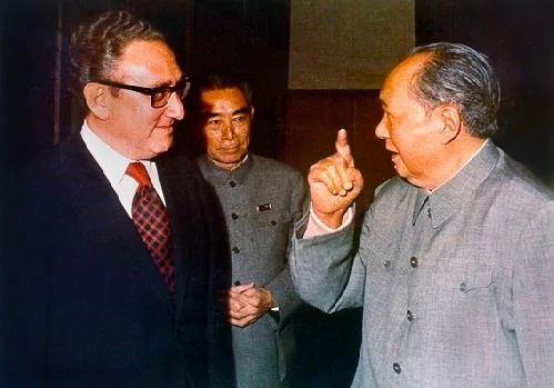 Henry Kissinger is depicted in a formal setting with a Chinese leader, both dressed in suits, signifying a diplomatic meeting. Kissinger is wearing a dark suit with a red and black patterned tie, exuding a serious and attentive demeanor. The Chinese leader, in a traditional Mao suit, is gesturing expressively with his hand, possibly emphasizing a point in conversation. The presence of another individual in the background, also in a Mao suit, suggests the importance of this meeting, potentially involving high-level discussions.
The setting is indoors, with dim lighting focusing on the individuals, creating a somber yet significant atmosphere. The attire and the respectful postures of the figures highlight the formality and importance of the encounter. The image captures a moment of international diplomacy, reflecting the political climate of the time.