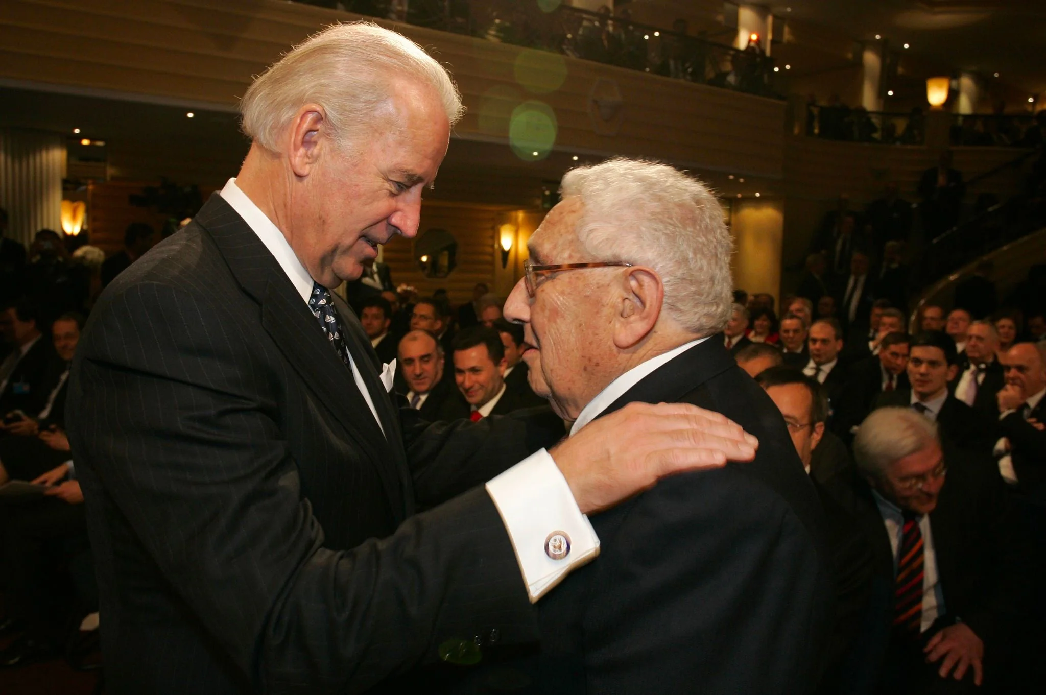 ```
In this image, Joe Biden is seen interacting with Henry Kissinger at what appears to be a formal event. The setting is indoors, with a large audience seated in the background. Joe Biden, on the left, is wearing a dark pinstripe suit with a white shirt and patterned tie. He has his hand on Kissinger's shoulder, demonstrating a friendly and respectful interaction. Henry Kissinger, on the right, is dressed in a dark suit and glasses, facing Biden closely. The atmosphere is formal, with attendees in suits and ties, suggesting a significant diplomatic or political gathering. The lighting is warm, highlighting their expressions and the cordial nature of the exchange. The audience is engaged, watching the interaction, adding to the sense of importance of the event.