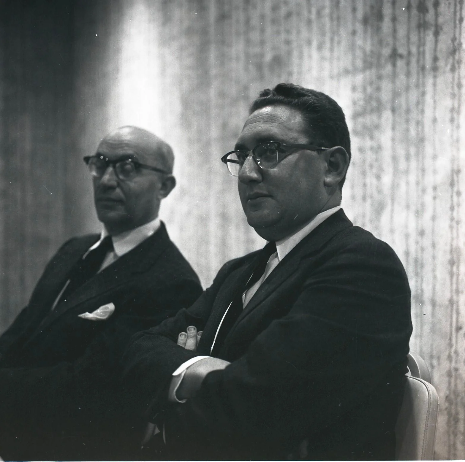 ```
The black and white image depicts two men seated side by side, both dressed in formal business attire. The man on the right is identified as Henry Kissinger, wearing a dark suit, a white shirt, and a striped tie. He is sitting with his arms crossed, appearing thoughtful and attentive. His glasses reflect the lighting, which is subtle and creates a soft contrast against the textured wall backdrop. The man on the left is also dressed in a suit and tie, with a pocket square visible in his jacket. Both men have a serious demeanor, suggesting a formal or professional setting, possibly a meeting or conference. The grayscale tone of the photograph enhances the classic and timeless feel of the scene. The focus is sharp on Kissinger, while the background and the other man are slightly blurred, highlighting the central figure's prominence in the image.