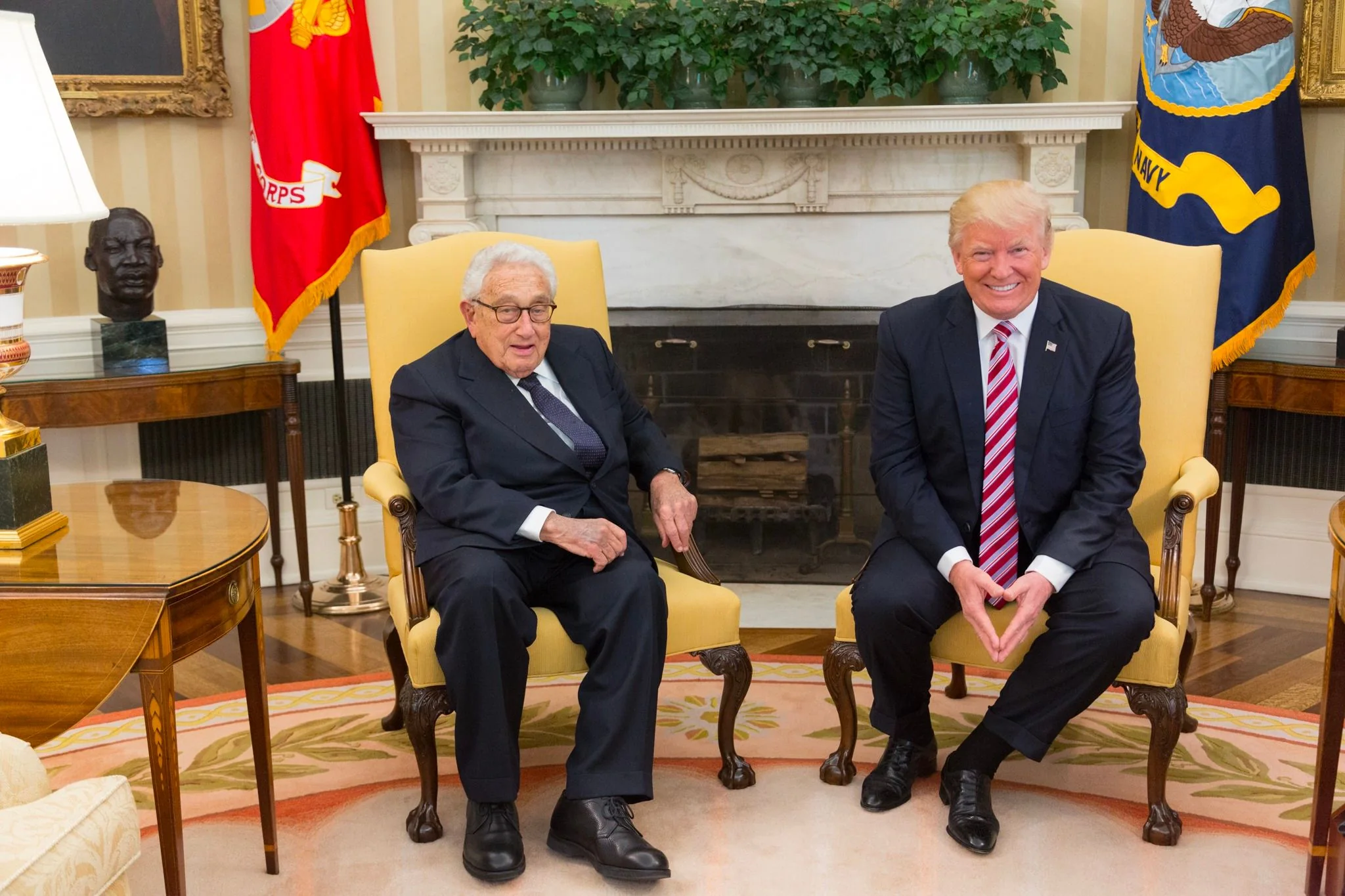 In this image, Henry Kissinger and Donald Trump are seated in a formal setting, likely an official meeting room. They are both sitting in elegant, yellow armchairs with wooden frames, positioned in front of a fireplace. Henry Kissinger is on the left, wearing a dark suit, white shirt, and a dark tie. He has a relaxed posture, with one hand resting on the armrest. Donald Trump is on the right, dressed in a dark suit, white shirt, and a red striped tie. He is sitting upright with his hands clasped. Both are smiling, suggesting a cordial interaction.
The room is richly decorated with a classic and formal aesthetic. Behind them, flags are displayed on either side of the fireplace, representing the Marine Corps and the Navy. Above the fireplace is a row of potted plants, adding a touch of greenery. The walls have vertical beige and white stripes, and a bust is placed on a side table to the left of Kissinger. The lighting is bright, emphasizing the formal and welcoming atmosphere of the room.