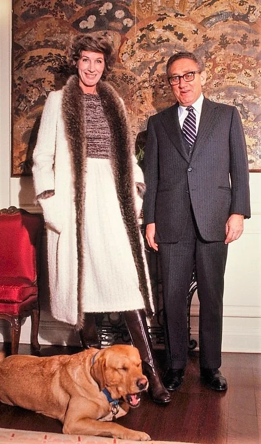 Henry Kissinger is pictured in a formal indoor setting. He stands to the right, dressed in a dark pinstripe suit with a white shirt and a striped tie. His posture is upright and he is smiling slightly at the camera. Beside him is a woman in a stylish outfit, wearing a white coat with a fur trim over a patterned dress and tall brown boots. They are positioned in front of an ornate, tapestry-like backdrop featuring intricate designs and patterns, adding a rich texture to the scene.
In the foreground, a golden retriever is lying on the floor, yawning or resting comfortably, adding a casual and warm element to the otherwise formal atmosphere. The room is elegantly furnished, with a red upholstered chair partially visible on the left. The lighting is soft yet clear, giving the scene a warm and inviting feel, emphasizing the luxurious and sophisticated ambiance.