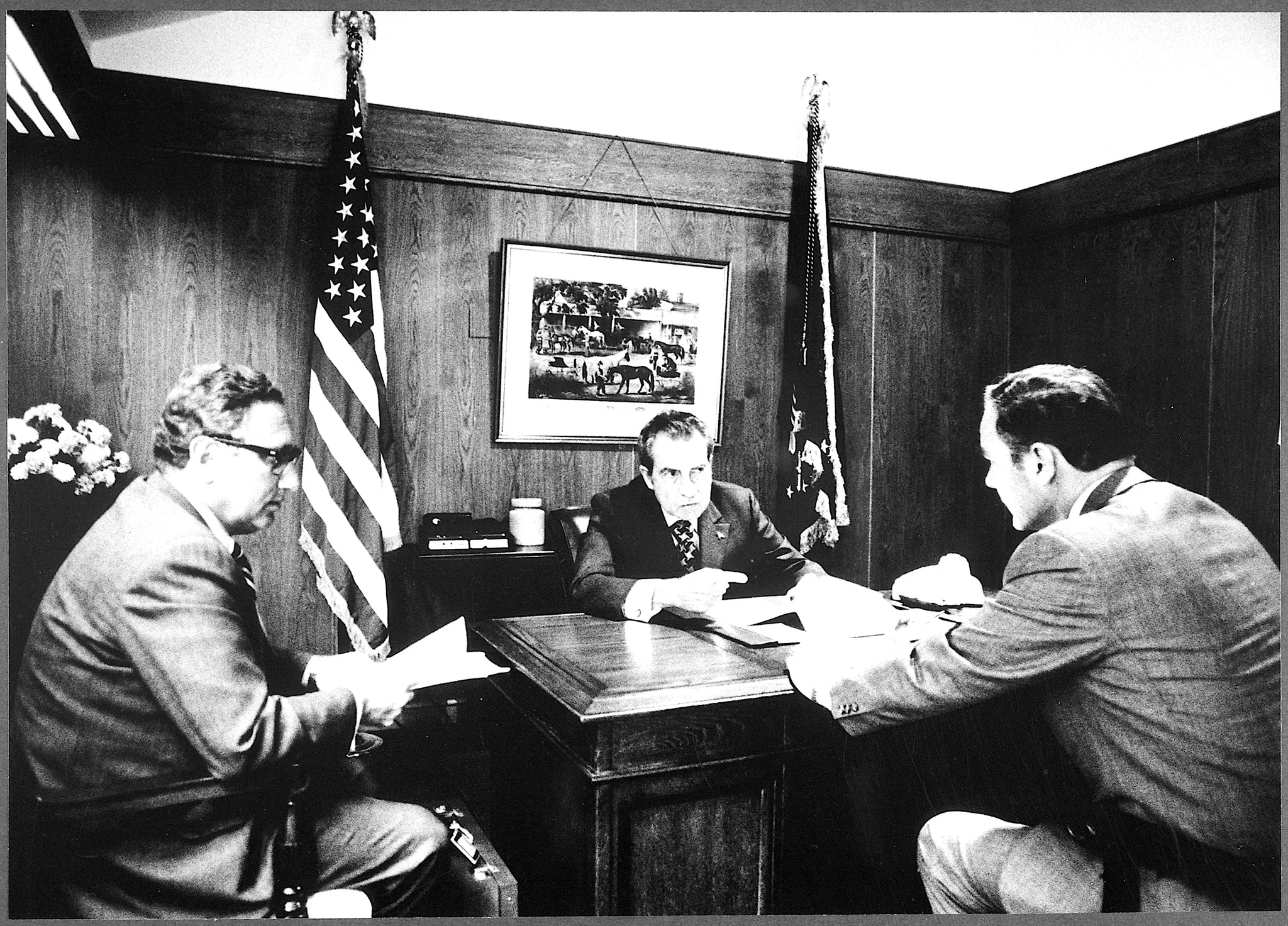 In this historical black and white image, three men are engaged in a meeting in a wood-paneled office. The man on the left, identified as Henry Kissinger, is seated, holding papers and wearing a suit and glasses. He appears to be listening intently. In the center, another man sits behind a wooden desk with an assertive posture, reviewing a document. Behind him, the American flag and another flag are prominently displayed, suggesting an official or governmental setting. A framed picture hangs on the wall above him, adding to the office's decor. The man on the right leans forward, actively participating in the discussion. The room is well-lit, emphasizing the formal and serious nature of the meeting. The overall mood is tense and focused, indicative of a significant political discussion or decision-making process.