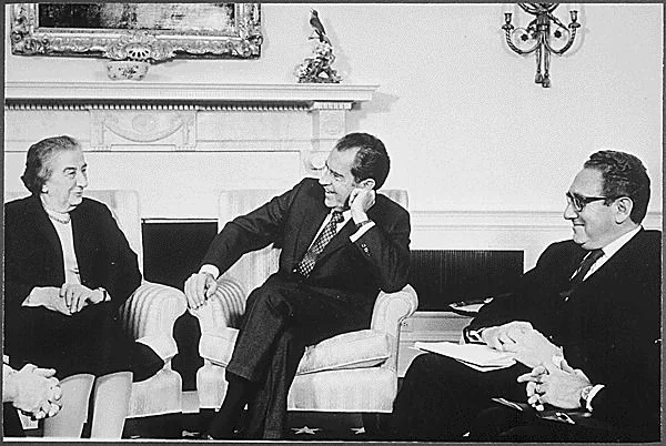 ```
In this historical black and white photograph, three individuals are seated comfortably in a formal room, likely a government office. The setting suggests a diplomatic meeting. On the left is a woman wearing a light-colored dress or suit, her hair styled traditionally, suggesting a formal occasion. In the center is a man, recognized from the hint as Nixon, wearing a dark suit and tie, sitting in a relaxed posture, leaning slightly towards the woman. He appears engaged in conversation, as indicated by his open expression and hand gesture. To the right is another man, identified as Henry Kissinger, also in a dark suit, holding papers, possibly notes or documents. The room’s decor is elegant, with an ornate fireplace in the background, adorned with a decorative piece and a painting above. The mood is professional yet cordial, suggesting a meeting of significance.