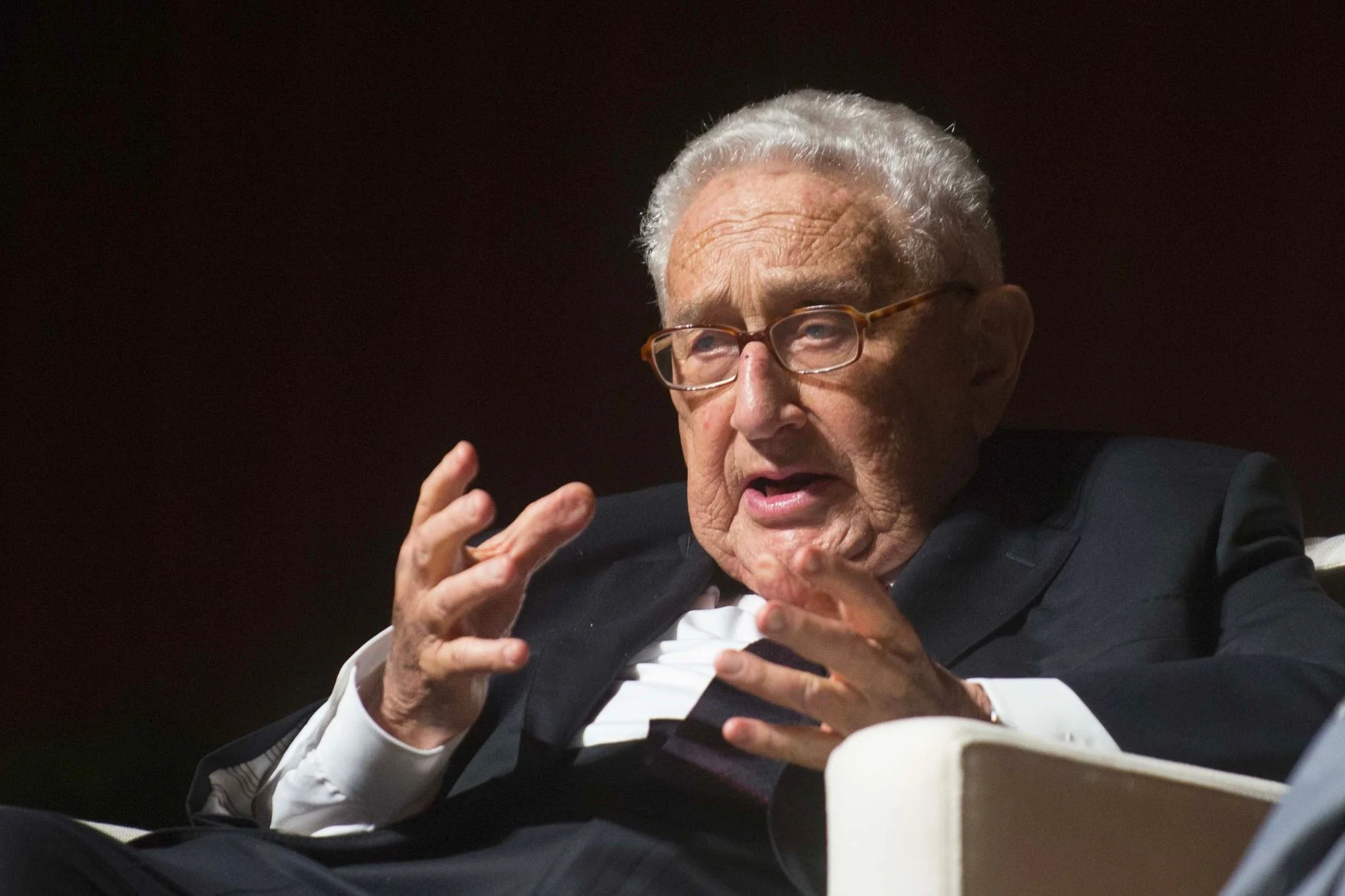 In the image, Henry Kissinger is seated on a stage, engaged in what appears to be a discussion or interview. He is gesturing with his hands, suggesting he is making a point or explaining something. Kissinger is wearing a dark suit, white shirt, and a dark tie, which adds to the formal atmosphere of the event. His expression is animated and thoughtful, indicating he is deeply engaged in the topic at hand.
The background is dark, which contrasts with the well-lit focus on Kissinger, drawing attention to him as the central figure. The lighting highlights his face and hands, emphasizing his expressions and gestures. The setting suggests a professional or academic event, possibly a symposium or conference. The overall composition conveys a sense of gravity and importance, fitting for a figure often associated with diplomatic and political discussions.