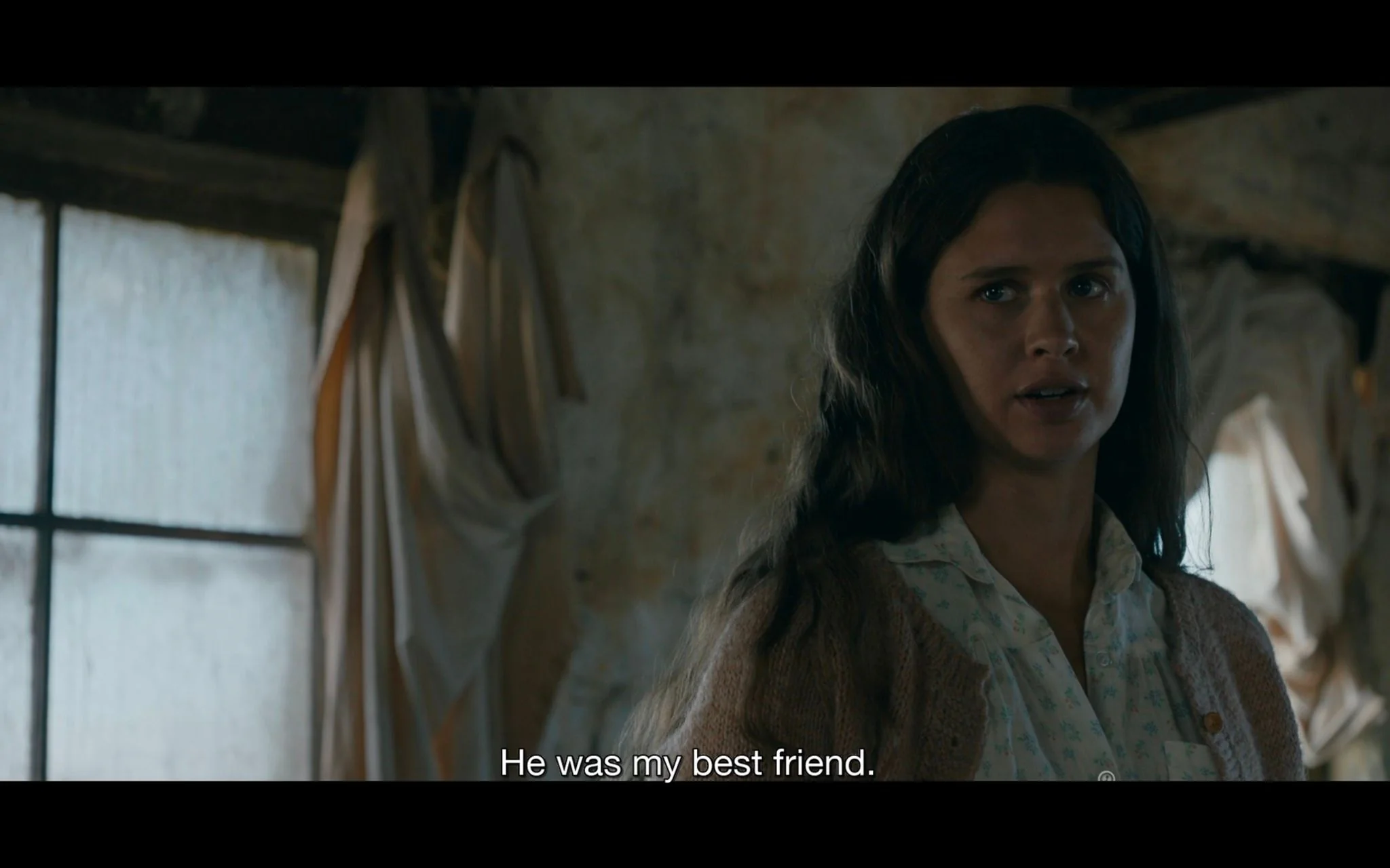 The image is a screenshot from the TV show "from," produced by MGM. It features a young woman indoors, likely part of a dramatic scene. She is positioned slightly off-center, looking towards someone or something outside the frame with a serious expression. Her long, dark hair is naturally styled, and she wears a light-colored blouse with a patterned cardigan. The background shows a rustic, somewhat worn interior with a window partially covered by draped fabric. The lighting is soft but dim, creating an intimate and somber atmosphere. The subtitle at the bottom of the image reads, "He was my best friend," indicating an emotional context. The scene suggests a moment of reflection or revelation, enhanced by the muted colors and the character's earnest demeanor.