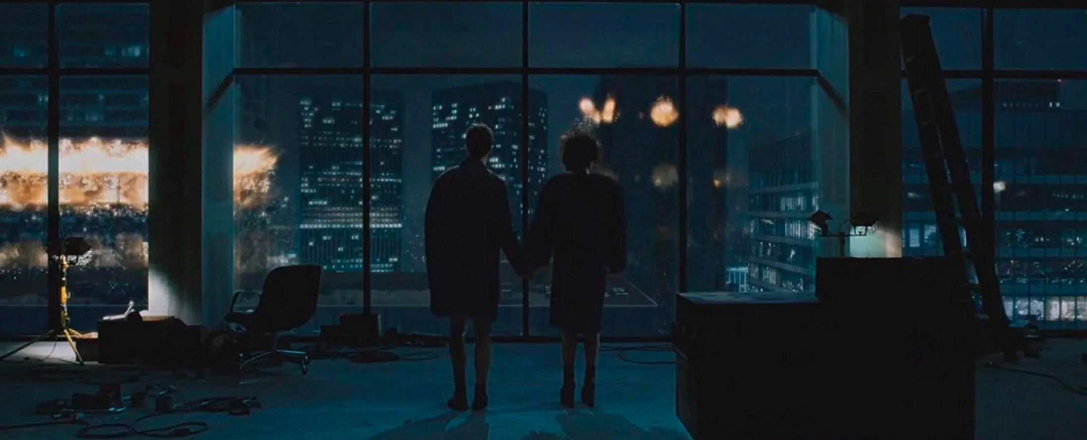 The image depicts a dramatic scene from the film "Fight Club." Two figures stand in the foreground, silhouetted against a large floor-to-ceiling window that reveals a nighttime cityscape. They are holding hands, conveying a sense of connection amidst chaos. The room is dimly lit, casting a bluish hue over the interior. Scattered objects on the floor and a ladder leaning against the wall suggest a setting of disarray and transition. Outside, buildings are illuminated with fiery explosions, creating a stark contrast between the calm interior and the tumultuous scene outside. The overall mood is tense yet intimate, with the figures seemingly unaffected by the destruction occurring beyond the glass. The composition of the shot draws the viewer’s eye toward the window, emphasizing the juxtaposition of personal and external conflict.