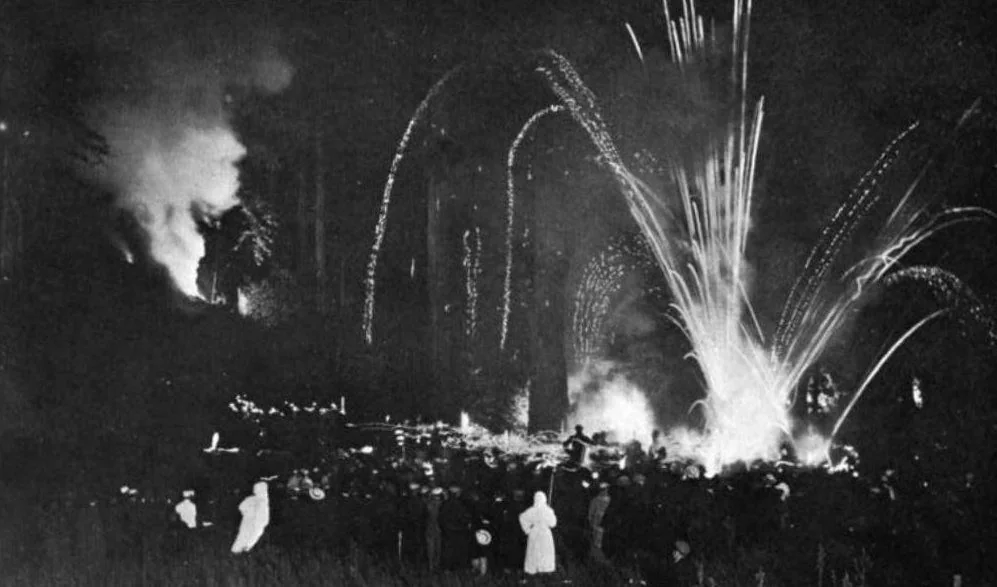 The image depicts a nighttime scene where a crowd of people is gathered outdoors, likely in a forested area, observing fireworks. The sky is illuminated with bright bursts of light, creating a dynamic display against the dark backdrop. In the foreground, the crowd appears engaged, with some individuals wearing light-colored clothing, standing out against the darkness. The atmosphere conveys a sense of celebration and festivity. The lighting primarily comes from the fireworks and possibly a large fire in the background, casting dramatic shadows and highlighting the outlines of the trees. The composition captures the vibrancy of the event, with the fireworks appearing as bright streaks arcing through the night sky.