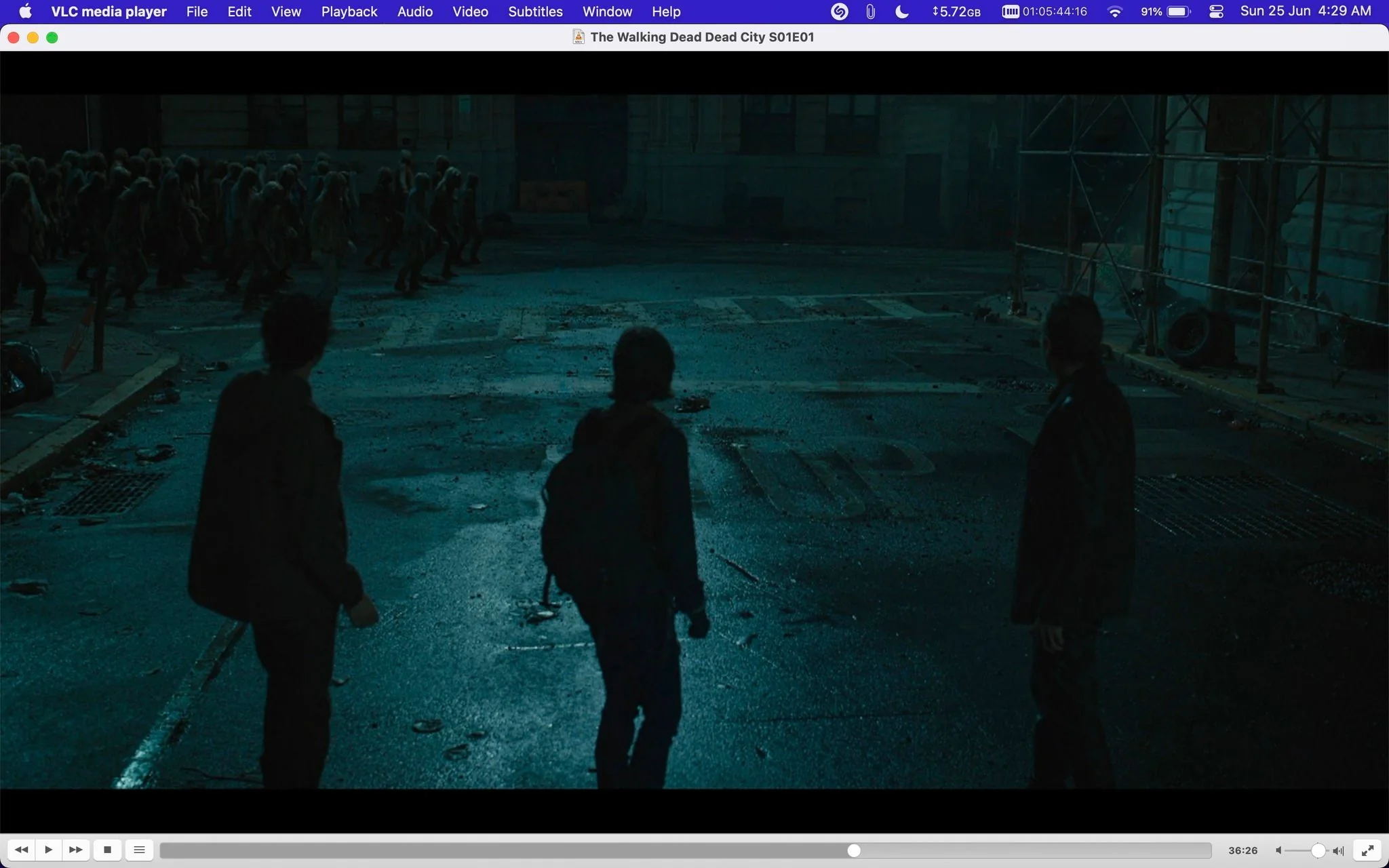 The image is a screenshot from the show "The Walking Dead Dead City" (Season 1, Episode 1) displayed on VLC media player. The scene takes place at night on an urban street, dimly lit with a bluish tint that highlights the tense atmosphere. In the foreground, three figures stand silhouetted against the street, observing a large group of zombies approaching from the background. The street is cluttered with debris, suggestive of a post-apocalyptic setting. The figures appear cautious, likely strategizing or preparing to confront the approaching threat. The lighting creates deep shadows, adding to the suspenseful mood. The urban environment, marked by building scaffolding and scattered litter, reinforces the desolate and dangerous world typical of "The Walking Dead" series.