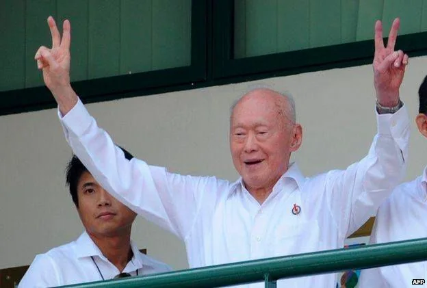 Lee Kuan Yew is featured prominently in this image. He is outdoors, standing on a platform or balcony, and is captured making a celebratory gesture with both hands raised in a "V" sign. Lee Kuan Yew is wearing a crisp white shirt adorned with a small pin on the left side, which might signify a political or organizational affiliation. His expression is joyful, indicating a moment of celebration or triumph. Next to him, a younger man is visible, also dressed in a white shirt, reflecting a sense of unity or team affiliation. The background features green panels, suggesting a structured setting, possibly a public or political event. The lighting is bright and natural, indicating a daytime setting. The image is credited to AFP, a known news agency, suggesting the photograph was taken during a significant public appearance.