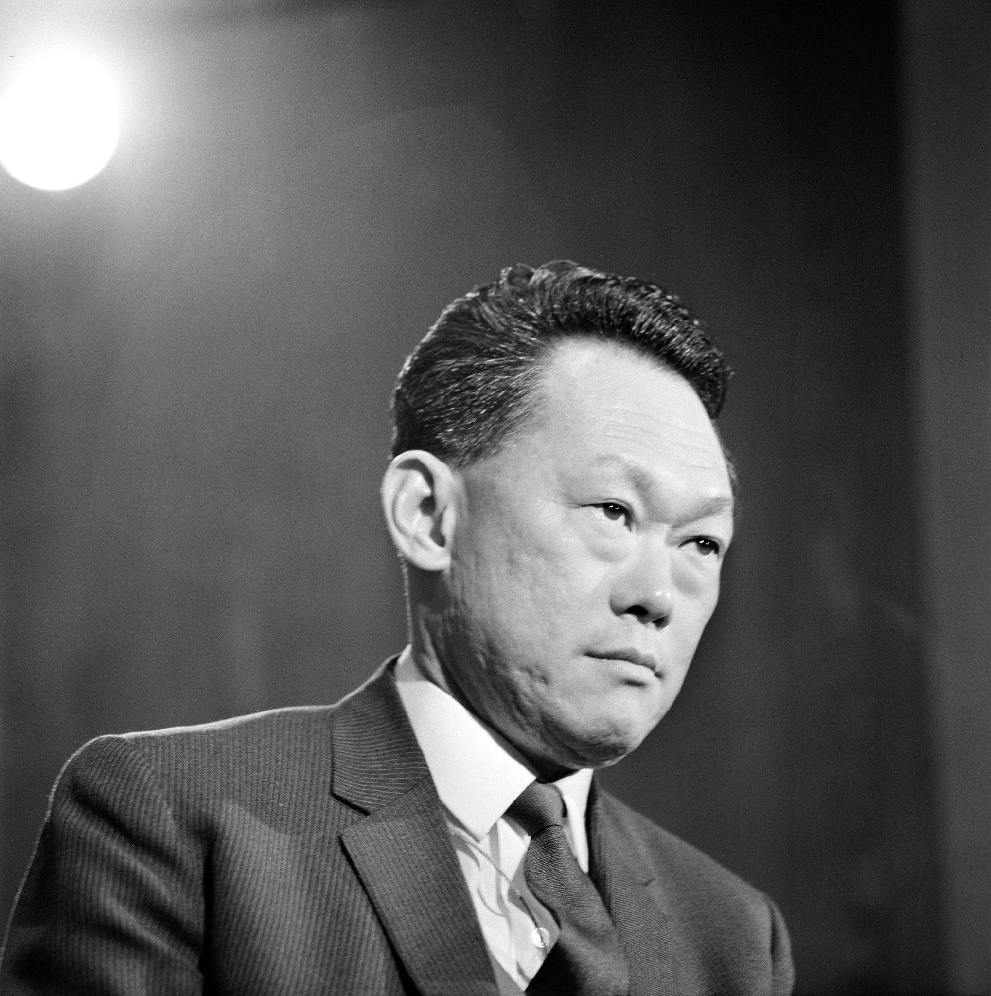 The image is a black and white portrait of Lee Kuan Yew, capturing a formal and contemplative moment. He is dressed in a suit and tie, signifying a professional or political setting. The lighting is focused on his face, creating a sharp contrast against the darker background, which enhances the serious expression on his face. His hair is neatly combed back, adding to the composed and authoritative presence he exudes. The absence of color emphasizes the historical and timeless nature of the photograph. The setting appears to be indoors, possibly during a formal event or interview, suggested by the spotlight illuminating him. The overall mood of the image is solemn and reflective, fitting for a leader of significant stature.