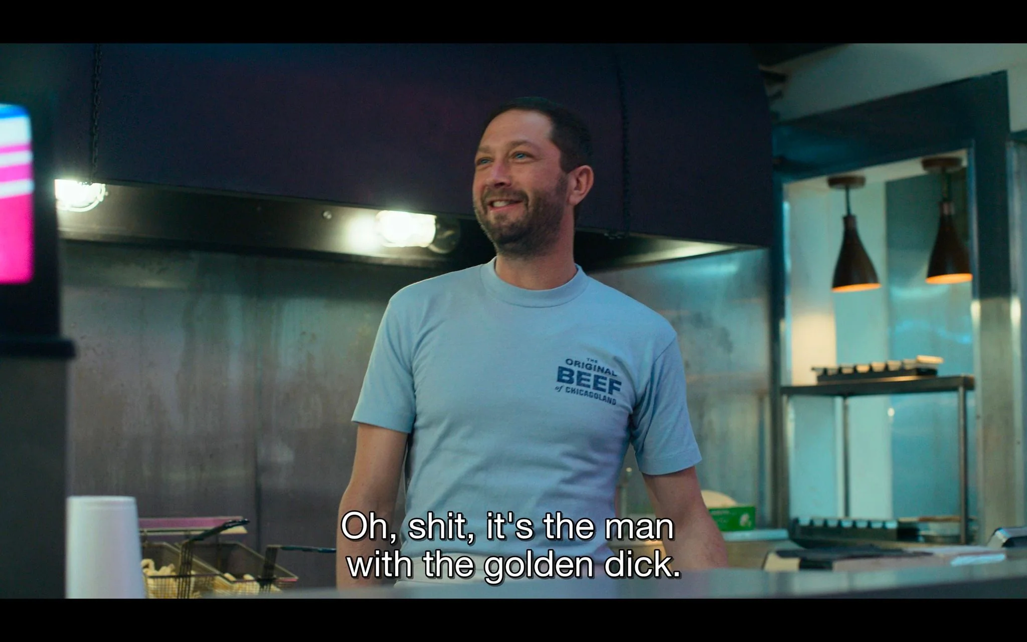 The image depicts a scene inside a restaurant kitchen, suggesting a casual and humorous setting. A man is standing, smiling slightly, and wearing a light blue T-shirt that reads, "THE ORIGINAL BEEF of CHICAGOLAND." This text and his presence in what appears to be a kitchen indicate that he might work there. The lighting is bright, highlighting the stainless-steel surfaces typically found in commercial kitchens. In the background, there are industrial kitchen fixtures, including lamps and shelves. The atmosphere seems lively and informal, emphasized by the playful caption, "Oh, shit, it's the man with the golden dick." This subtitle adds a comedic element to the scene. The man’s relaxed expression and posture contribute to the laid-back and friendly mood of the image.