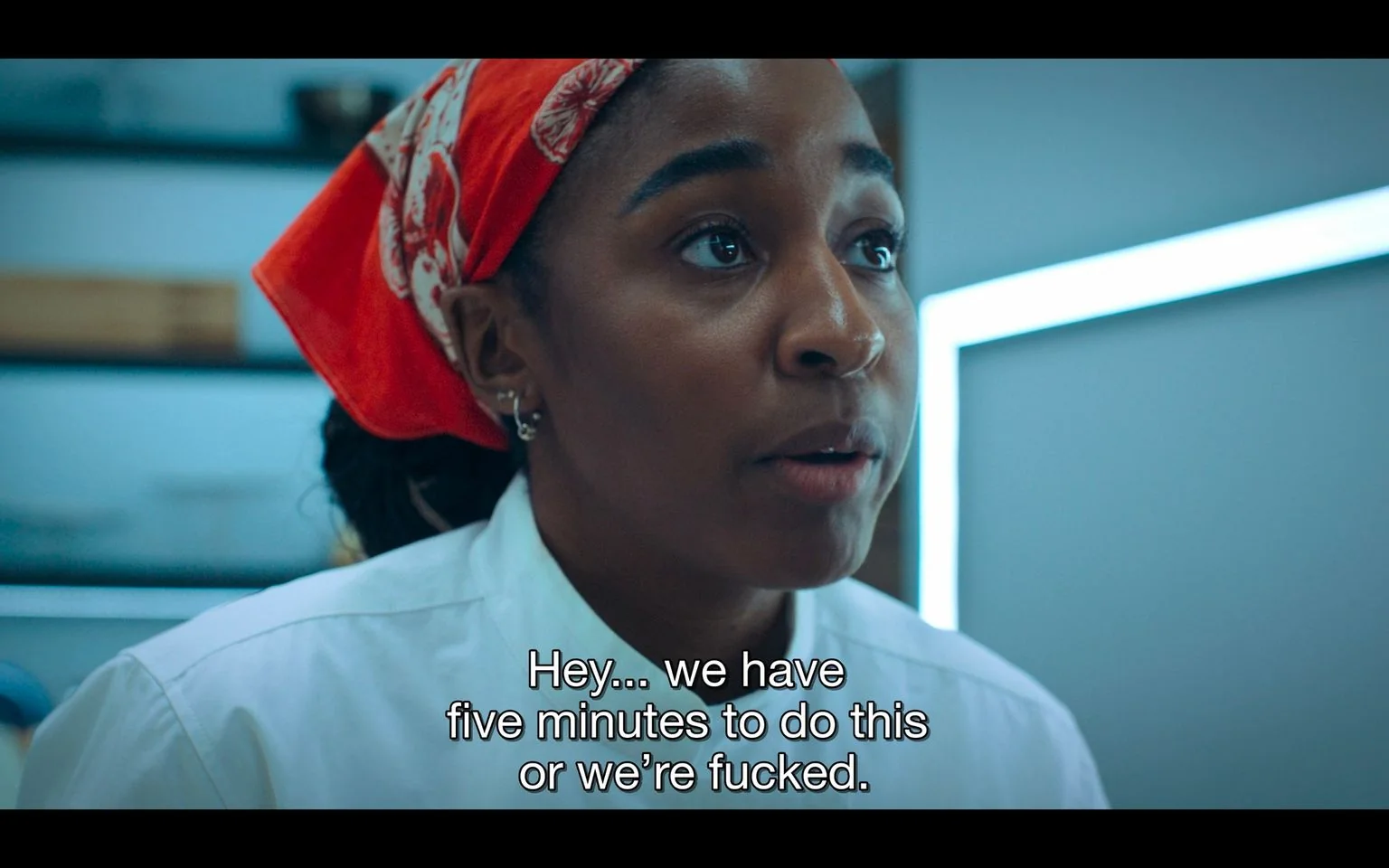 The image shows a close-up of a woman in a kitchen setting. She is wearing a white chef’s jacket and a red bandana with white patterns, suggesting that she is a chef or cook. Her expression is serious and focused, indicating urgency or stress. The background is blurred, but the setting appears to be a modern kitchen with bright, cool-toned lighting. The woman seems to be speaking to someone off-camera, as indicated by the subtitle: "Hey... we have five minutes to do this or we’re fucked." The subtitle suggests a tense or high-pressure situation, possibly related to cooking or preparing a dish in a limited time frame. The composition focuses on her face, emphasizing her expression and the urgency of the moment.