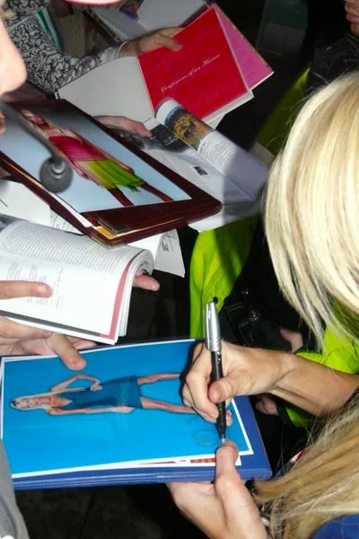 The image captures a busy scene of autograph signing, likely at an event where fans gather to meet a public figure. In the foreground, a person with blonde hair is holding a pen and signing a photograph for someone. The photograph being signed features an individual in a blue outfit, and multiple hands are visible, holding various items like books and magazines, suggesting a crowd eager for autographs. The setting appears to be indoors with a bustling atmosphere. The person signing is dressed casually, with their focus on the task, indicating a sense of engagement with the fans. The lighting is bright, illuminating the scene and highlighting the colorful items being held by the crowd. There is a sense of excitement and anticipation among the people present, typical of such fan events.