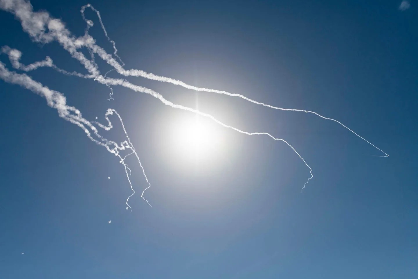 The image captures a clear blue sky with the sun shining brightly, creating a dramatic and vivid scene. Several white smoke trails curve and twist across the sky, likely indicating the paths of aircraft or missiles. The sun, positioned centrally, casts a strong light, illuminating the trails and enhancing their contrast against the blue background. The composition suggests motion, with the trails diverging and curving in different directions. This visual is dynamic, suggesting a scene of movement or activity, possibly related to an air show or military exercise. The simplicity of the elements—primarily the sky and the smoke—focuses attention on the patterns and the play of light. The overall mood is one of energy and clarity, with the bright sunlight adding to the intensity of the scene.