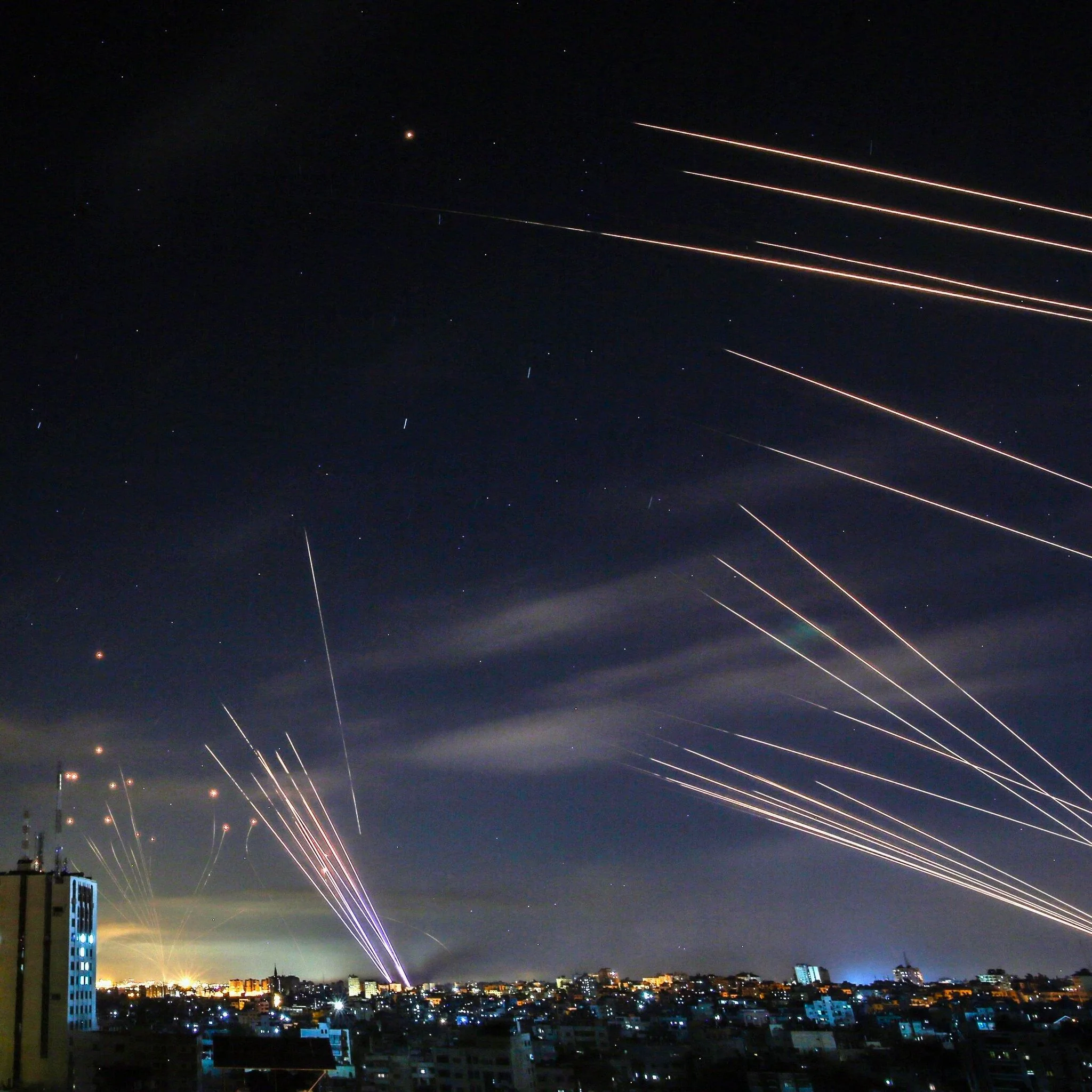 This image captures a dramatic night scene over a city landscape. The foreground shows a densely built urban area, illuminated by artificial lights. The skyline is dotted with buildings, some of which are taller, featuring antennas or spires. The background is dominated by the night sky, which is alive with streaks of light, likely captured using long exposure photography. These streaks suggest the launch or flight of rockets or missiles, creating an intense and dynamic visual.
The sky appears mostly clear, with some light clouds and stars visible, adding depth to the scene. The colors in the image range from the warm, orange hues of city lights to the cooler, bluish tones of the night sky. The overall mood is tense and dramatic, with the trajectory of the streaks indicating motion and urgency. The composition effectively contrasts the stillness of the city below with the dynamic action in the sky above, creating a visually striking image.