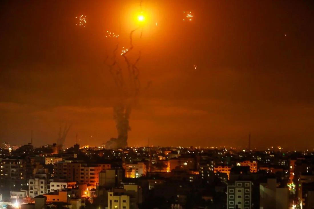 A dramatic night scene unfolds over a city, with buildings silhouetted against the glow of multiple explosions or fireworks in the sky. The image captures a large, bright orange burst at the center, surrounded by smaller sparkling lights and trails of smoke. The skyline is densely packed with buildings, their windows lit against the darkness, suggesting a populated urban area. The sky is dominated by the vibrant orange hue, likely from the explosion, casting an eerie glow over the scene. The trails leading to the explosions create a sense of motion and chaos, indicating a potentially intense or significant event.
The overall mood is tense and dramatic, with the lighting emphasizing the contrast between the illuminated sky and the shadowed city below. The lack of visible people or specific landmarks keeps the focus on the sky and the unfolding event. The scene is captured in a photographic style, suggesting it is a real-world event rather than a fictional or artistic depiction.