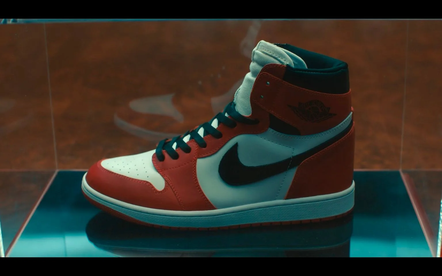 The image showcases a classic high-top sneaker, displayed in a transparent case likely designed for exhibition purposes. The sneaker's design prominently features a bold red, black, and white color scheme, characteristic of a popular and iconic brand. The shoe's upper is primarily white, with vivid red overlays on the toe, side panels, and heel. A striking black swoosh logo decorates the side, adding to the signature look. The shoe's laces are black, contrasting with the white tongue, which adds a clean, elegant touch to the overall design.
The background is blurred, focusing attention on the sneaker itself, which is positioned slightly to the left within the frame. The lighting is soft, creating subtle reflections on the glass and emphasizing the shoe's vibrant colors. The setting appears to be a showroom or a display area, with the sneaker's iconic design likely drawing the attention of enthusiasts and collectors. The mood is both nostalgic and contemporary, reflecting the lasting appeal of this footwear style.