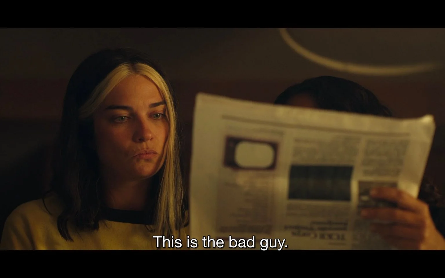 The image features a scene set indoors with dim lighting, creating an intimate and somewhat tense atmosphere. In the foreground, a person with distinctive hair—dark with prominent light streaks—sits with a focused yet pensive expression. They are wearing a yellow top, and their gaze is fixed intently on a newspaper, which is held up by another person partially obscured from view. The newspaper is in the center of the frame, suggesting it contains significant information pertinent to the scene. The caption at the bottom, "This is the bad guy," implies a moment of revelation or identification, possibly in a narrative context such as a film or TV show. The overall color palette is warm, with browns and yellows dominating the scene, enhancing the mood of quiet intensity.