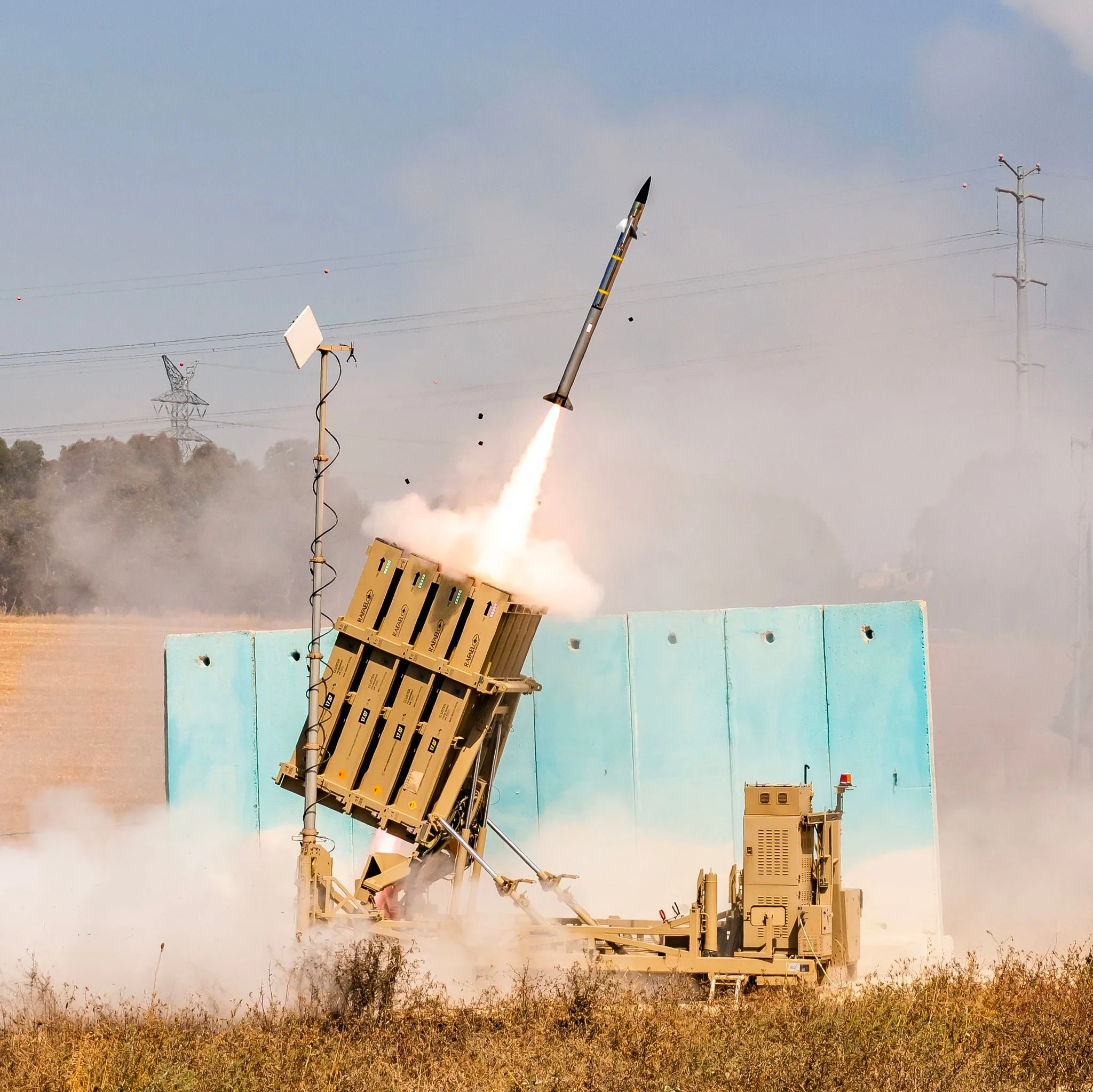 This image depicts a missile defense system in action, likely an Iron Dome battery, based on the setup and context. The scene captures a missile being launched into the sky, with a trail of smoke and fire indicating its rapid ascent. The launcher is angled slightly upward, positioned on a platform surrounded by a rugged outdoor environment typical for military installations. The sky is clear, with some power lines visible in the background, suggesting a rural or semi-rural setting.
The launcher consists of several missile containers, arranged in a neat configuration, reinforcing the organized and technical nature of military operations. The words "YAHALOM" are visible on the containers, indicating a specific model or component of the system. The scene conveys a sense of urgency and precision, typical of defense operations. The colors are muted, with the launcher and background primarily in shades of beige and green, blending into the natural surroundings. This setting is likely part of a demonstration or active defense scenario, showcasing the capability and readiness of the missile system.