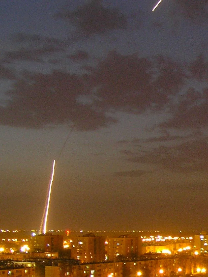 This image captures a striking night scene with a rocket launch captured in long exposure. The sky is dark, with scattered clouds creating a moody atmosphere. A bright streak of light arcs diagonally from the bottom left, where the rocket appears to have launched, heading towards the upper right, gradually fading out. This light trail indicates the path of the rocket as it ascends into the night sky. Below, the urban landscape is illuminated by the warm glow of streetlights, outlining several buildings in the foreground. The structures appear to be residential, with multiple windows visible, some lit up. The overall color palette includes deep blues of the night sky, contrasted by the warm yellows and oranges of the city lights, contributing to the dramatic effect of the scene.
