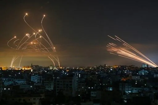 This image captures a dramatic nighttime scene over a city skyline. The foreground is filled with densely packed buildings, their outlines illuminated by streetlights and the glow of the city. In the sky above, a striking visual display unfolds: multiple streaks of light trace across the night sky. On the left side, several arcs of light are visible, likely representing the interception of projectiles in midair. These arcs are characteristic of an active defense system responding to incoming threats. On the right, a cluster of straight, bright trails suggests the launch or flight of rockets or missiles. The contrast between the warm glow of the city and the cold, stark lines of light in the sky creates a tense and dramatic atmosphere, highlighting the ongoing conflict and defense measures in place. The image is both compelling and somber, capturing the reality of modern warfare technology against a civilian backdrop.