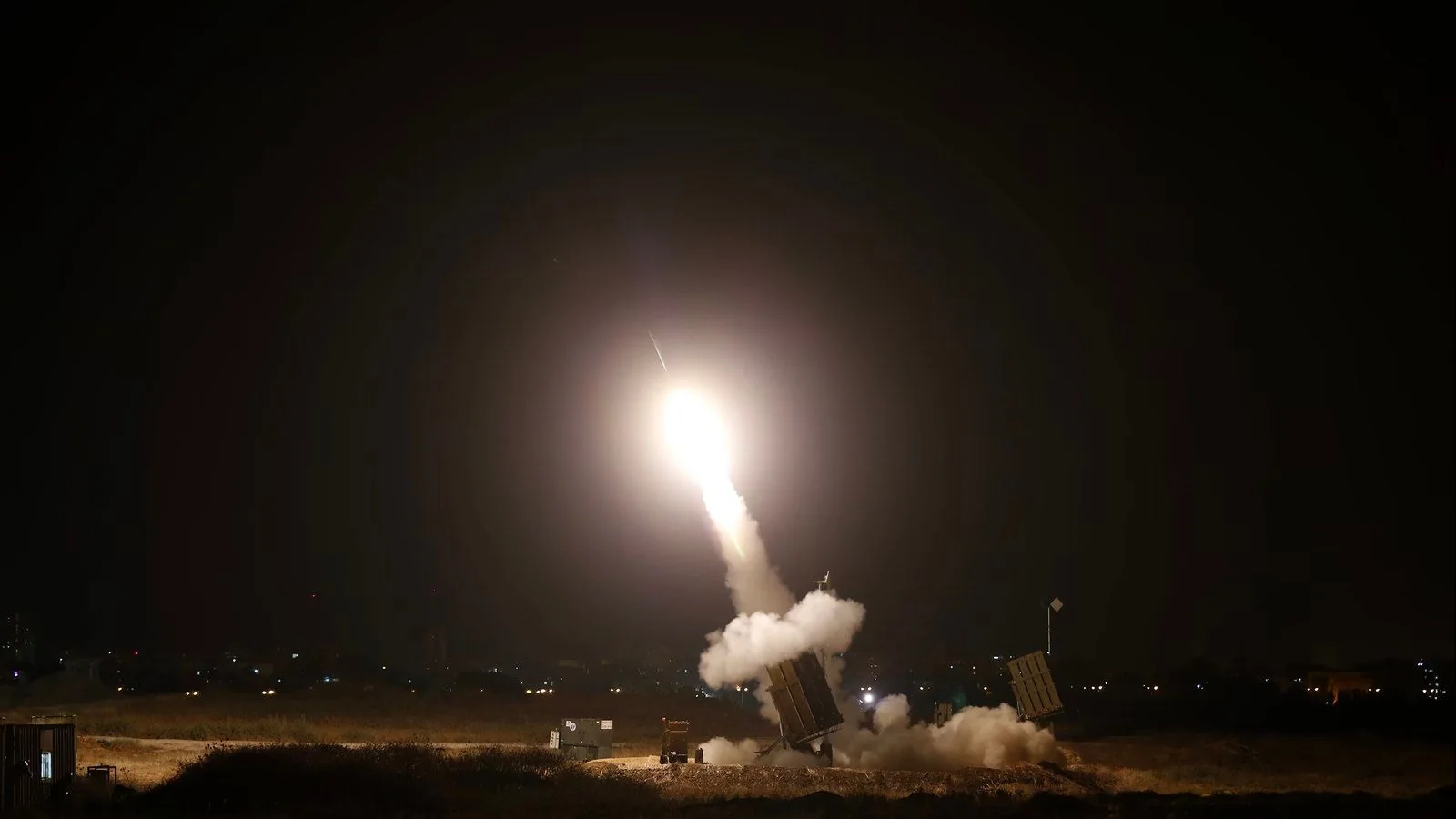 The image shows a night-time scene of a missile being launched from a military defense system setup. The environment is dark, with the launch creating a bright, fiery glow against the night sky. The missile is ascending at a steep angle, leaving a trail of smoke and light behind. The launcher is positioned on the ground, surrounded by dry grass, and emits a cloud of smoke as the missile is fired. In the background, the silhouette of a city skyline with scattered lights can be seen, suggesting the location might be near an urban area. The scene conveys a sense of urgency and precision, characteristic of military operations. The image captures the dramatic contrast between the bright launch and the dark surroundings, emphasizing the power and technology involved in the defense system.
