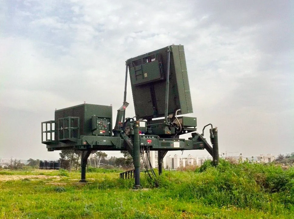 ```
The image showcases a military radar system situated in an open field with a grassy foreground. The equipment is elevated on sturdy legs, allowing for stability and elevation in uneven terrain. Its design includes a large rectangular antenna panel, suggesting it is used for tracking or detection purposes. The equipment is predominantly green, which is typical for military technology to blend with natural surroundings.
In the background, there are trees and distant buildings, indicating the location might be near an urban area. The sky is overcast, casting a diffuse light over the scene, which softens shadows and enhances the muted colors of the equipment and the natural environment. There are no visible people or additional vehicles, focusing the viewer's attention solely on the radar system. This setup likely represents part of a larger defense mechanism, possibly used for monitoring airspace or detecting incoming threats.