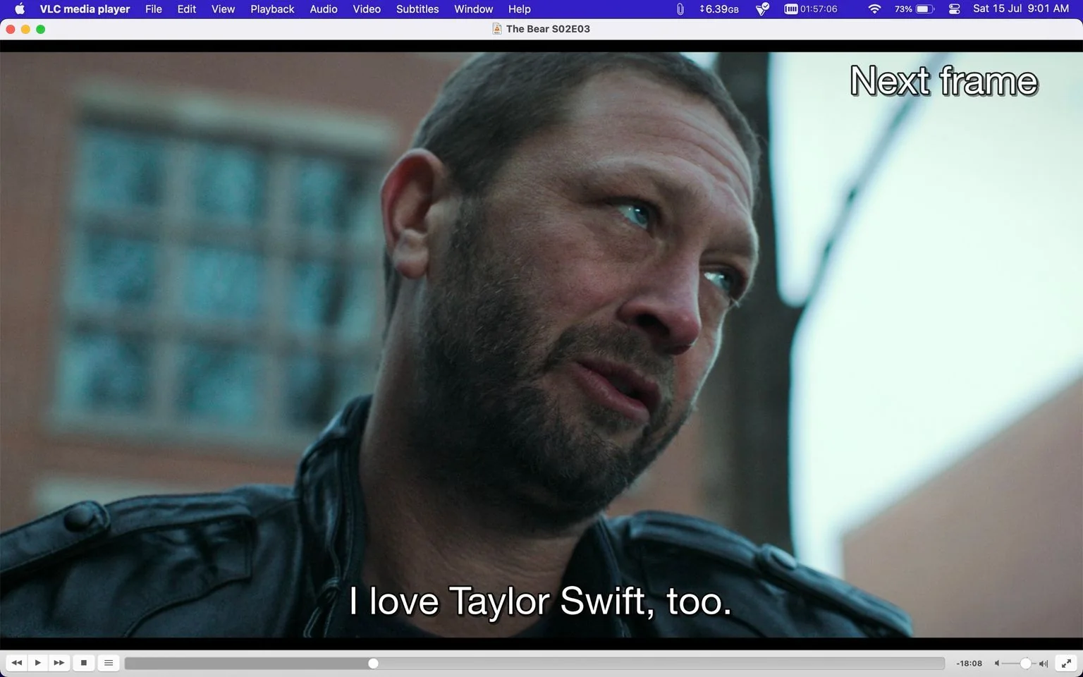 The image is a screenshot from the series "The Bear," specifically from Season 2, Episode 3. It is displayed on a computer screen using VLC media player, as indicated by the interface elements at the top and bottom of the image. The scene features Ebon Moss-Bachrach portraying the character Richard 'Richie' Jerimovich. He is outdoors, wearing a dark leather jacket, and his expression is contemplative as he looks slightly upwards. The background is blurred, showing a building and some trees, suggesting an urban environment. The lighting is natural, likely indicating daytime. The subtitle reads, "I love Taylor Swift, too," conveying a moment of personal expression or connection. The composition focuses on Richie, capturing his emotional state in a candid moment.