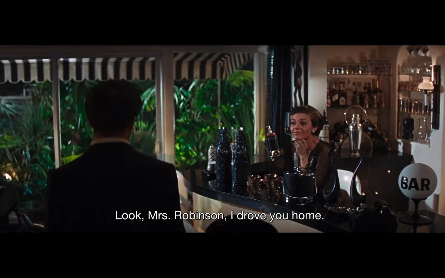 The image is a scene from the film "The Graduate." It shows a nighttime indoor setting, specifically inside a well-appointed home bar area. On the right, a woman identified as Mrs. Robinson is seated at the bar, looking contemplative and composed. She is elegantly dressed, suggesting sophistication and affluence. The lighting is soft, casting a warm ambiance over the scene. In the foreground, a man is seen from behind, addressing Mrs. Robinson, as indicated by the subtitle: "Look, Mrs. Robinson, I drove you home."
The bar is stylishly equipped with various bottles and glasses, and there's a "BAR" sign prominently displayed. Outside the windows, lush greenery is visible, adding to the upscale and private atmosphere. This scene likely captures a moment of tension or revelation, typical of the film's dramatic narrative. The composition emphasizes the dialogue between the characters, focusing on their interaction and the intimate setting.