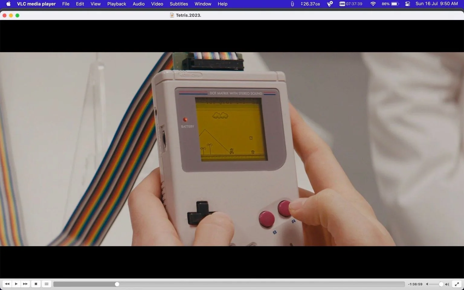 The image is a video frame from the movie titled "Tetris." It shows a close-up of a person holding a Game Boy, a classic handheld gaming console. The Game Boy's screen displays a simple game with pixelated graphics featuring clouds, pyramids, and palm trees, indicating a retro gaming theme. The person’s hands are visible, with fingers positioned on the directional pad and buttons, suggesting active gameplay. The Game Boy features distinct text at the top, “DOT MATRIX WITH STEREO SOUND,” and the “BATTERY” indicator is illuminated.
The background is out of focus, emphasizing the handheld console. A colorful ribbon cable is visible, adding a splash of color against the muted tones of the Game Boy. The lighting is bright and even, typical of indoor settings, providing clear visibility of the on-screen elements and console details. The use of the VLC media player interface suggests the image is taken from video playback on a computer, with controls and file information displayed at the top and bottom of the screen. The media file is named "Tetris.2023.1080p.WEBRip.x264.AAC5.1-[YTS.MX].mp4," indicating that this frame is from a new adaptation or documentary related to the game Tetris.