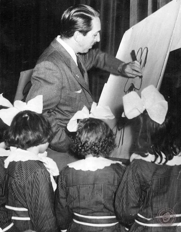 Walt Disney is captured in this black-and-white image, engaged in drawing on a large easel. The scene takes place indoors, with Walt Disney surrounded by a small group of children, who appear to be intently watching his artistic process. The children, all girls, are dressed similarly in striped dresses with large bows in their hair, suggesting a formal or school setting. Walt Disney is wearing a suit, portraying a professional yet approachable demeanor as he sketches. The drawing he is working on seems to depict a familiar character, recognizable by the large ears, which may be indicative of his iconic creations. The atmosphere is one of curiosity and learning, as the children seem captivated by the scene. The lighting is soft, focusing on the interaction between Walt Disney and the children. The composition emphasizes the connection between the artist and his young audience, reflecting a moment of creativity and inspiration.