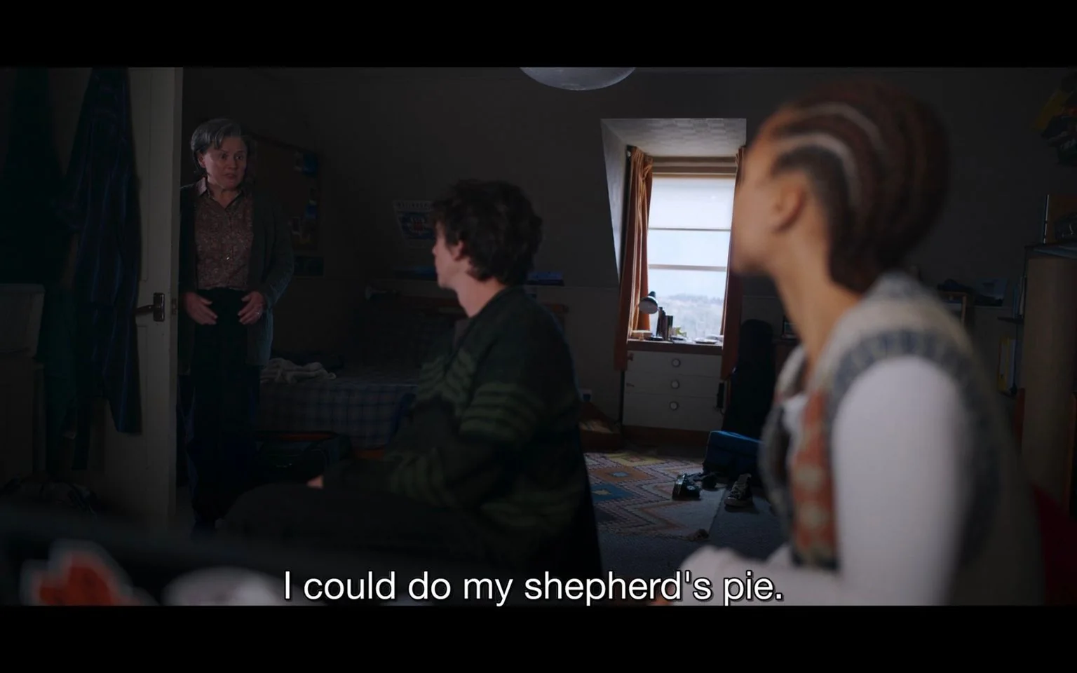 ```
The image depicts an indoor scene in a dimly lit bedroom. A woman stands at the door, wearing a patterned blouse and a cardigan, looking at two individuals seated in the room. Her expression appears inviting as she suggests, indicated by the subtitle, "I could do my shepherd's pie." One of the seated individuals, a man, is turned around to face her, while the other person, a woman with braided hair, is partially visible from the side. The room is cozy and slightly cluttered with a bed, a window with curtains drawn to the side, and a desk with various items, including a lamp and what appears to be books or papers. The setting suggests a casual and intimate atmosphere, likely a home environment where the conversation is taking place. The lighting is soft, creating a warm ambiance in the room.